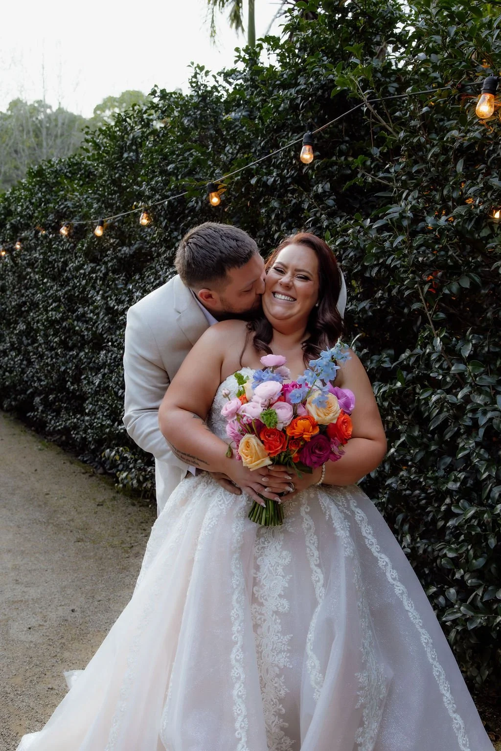 A newlywed couple smiling, with the groom kissing the bride's cheek, standing outdoors near a hedge with string lights overhead. The bride is holding a colorful bouquet of flowers.