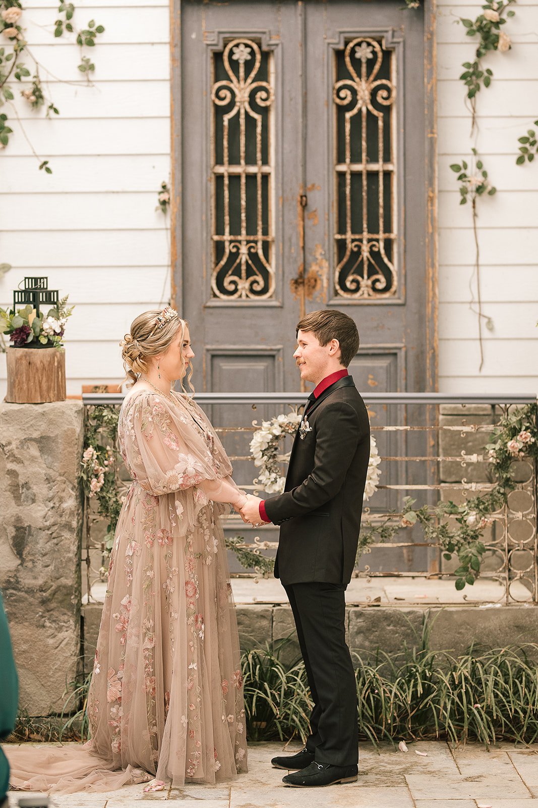 A couple holding hands and looking at each other during a wedding ceremony. The bride is wearing a floral embroidered gown and the groom a black suit with a red shirt, standing in front of a rustic wooden door with metal accents, decorated with flowers.