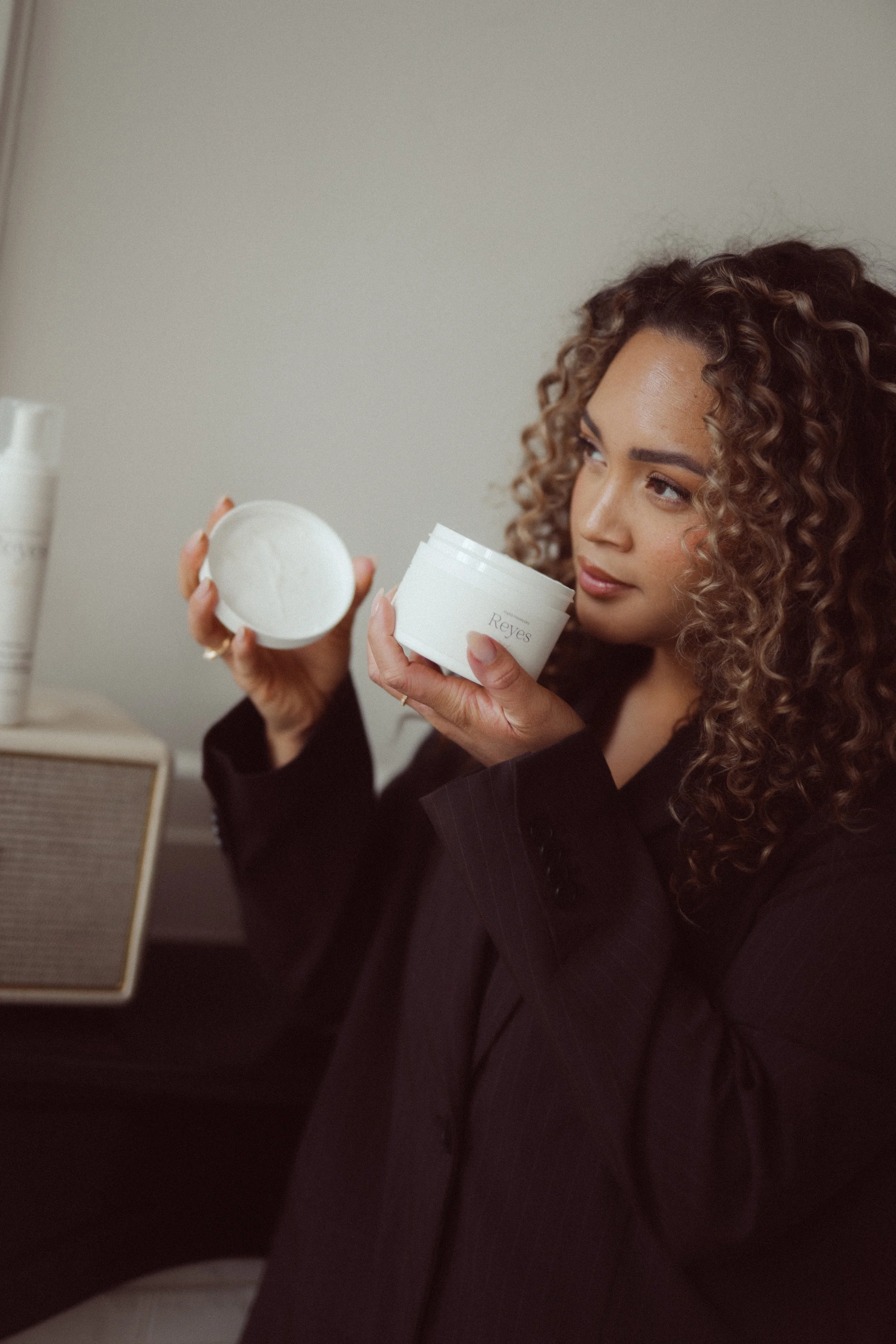 Een vrouw met krullend haar die een pot crème vasthoudt en naar de inhoud kijkt.