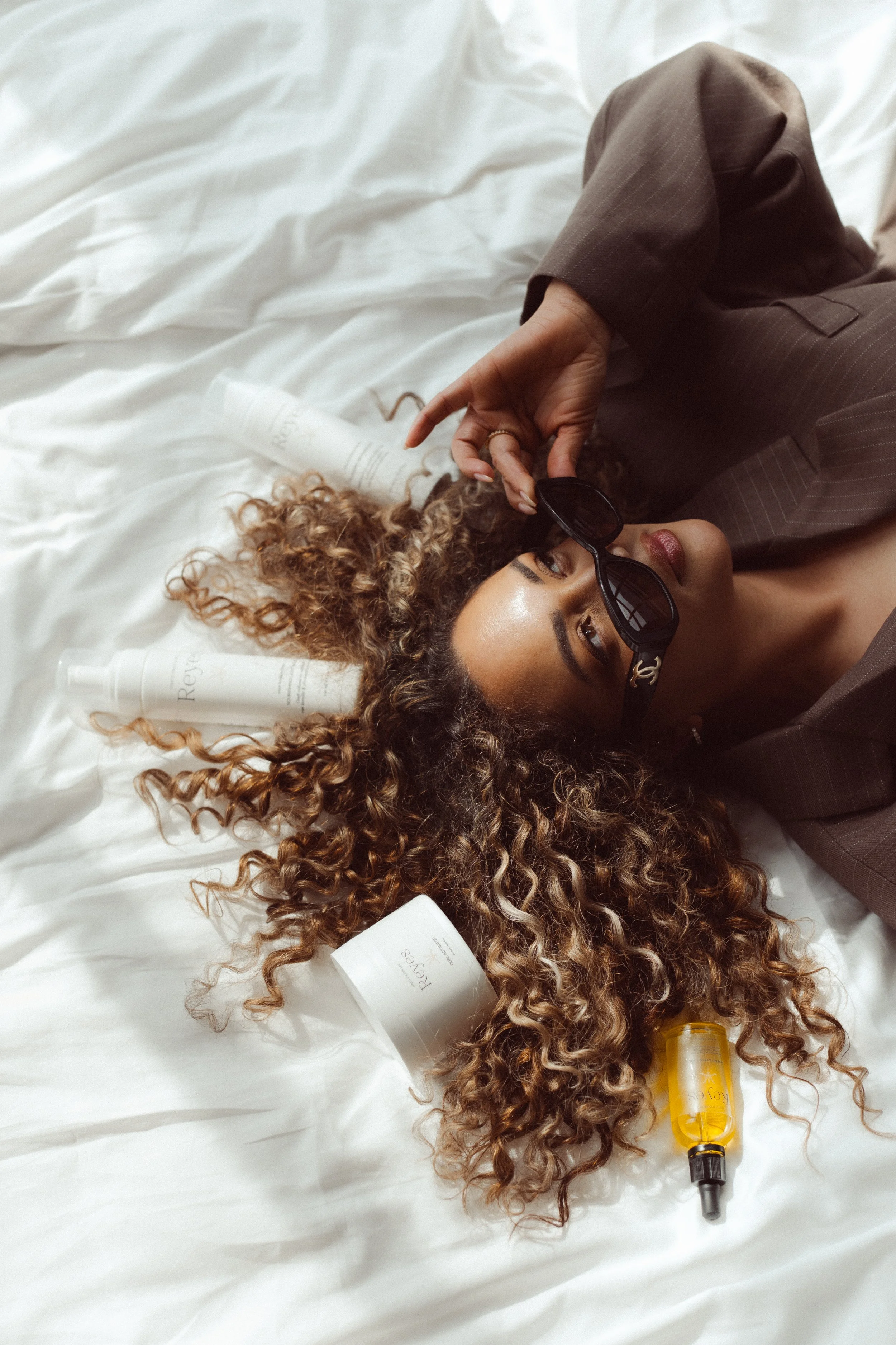 Een vrouw met gekrulde haar ligt op een wit bed, met zonnebril, omringd door haarverzorgingsproducten, zoals shampoo en haarolie.