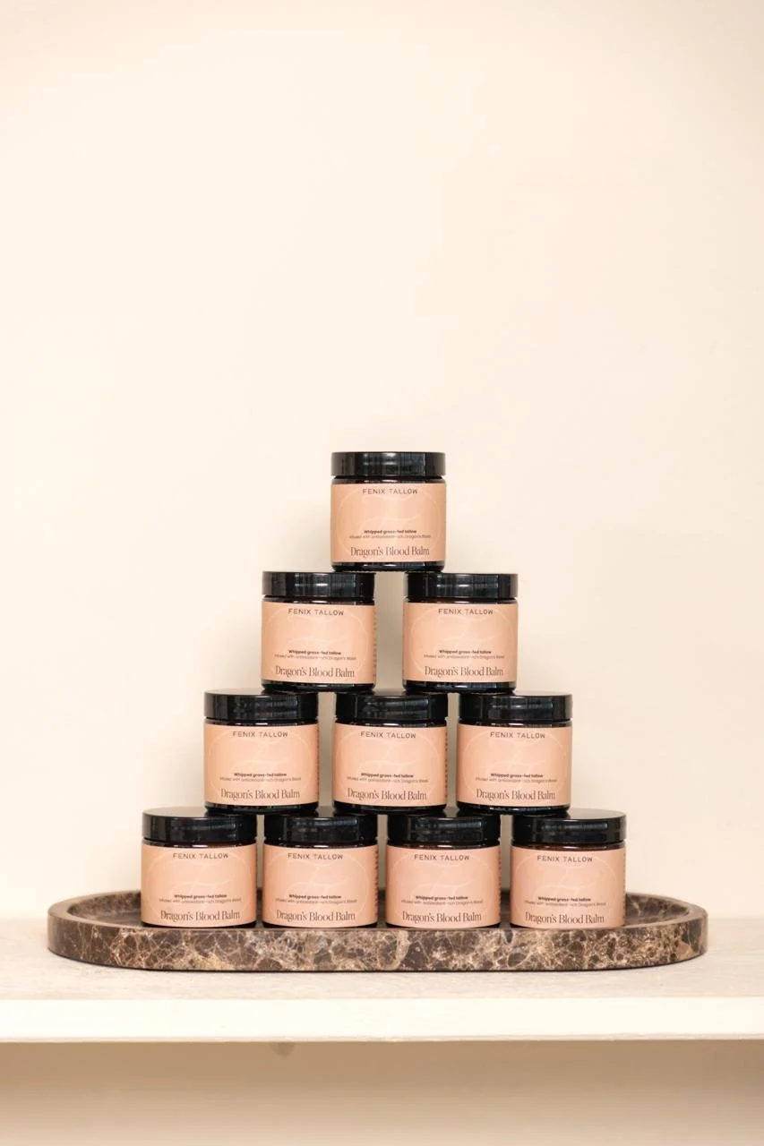 Tien potten met huidverzorgingsproduct, genaamd 'Dragon's Blood Balm' van Fenix Tallow, gestapeld op een ronde stenen schaal op een witte plank.