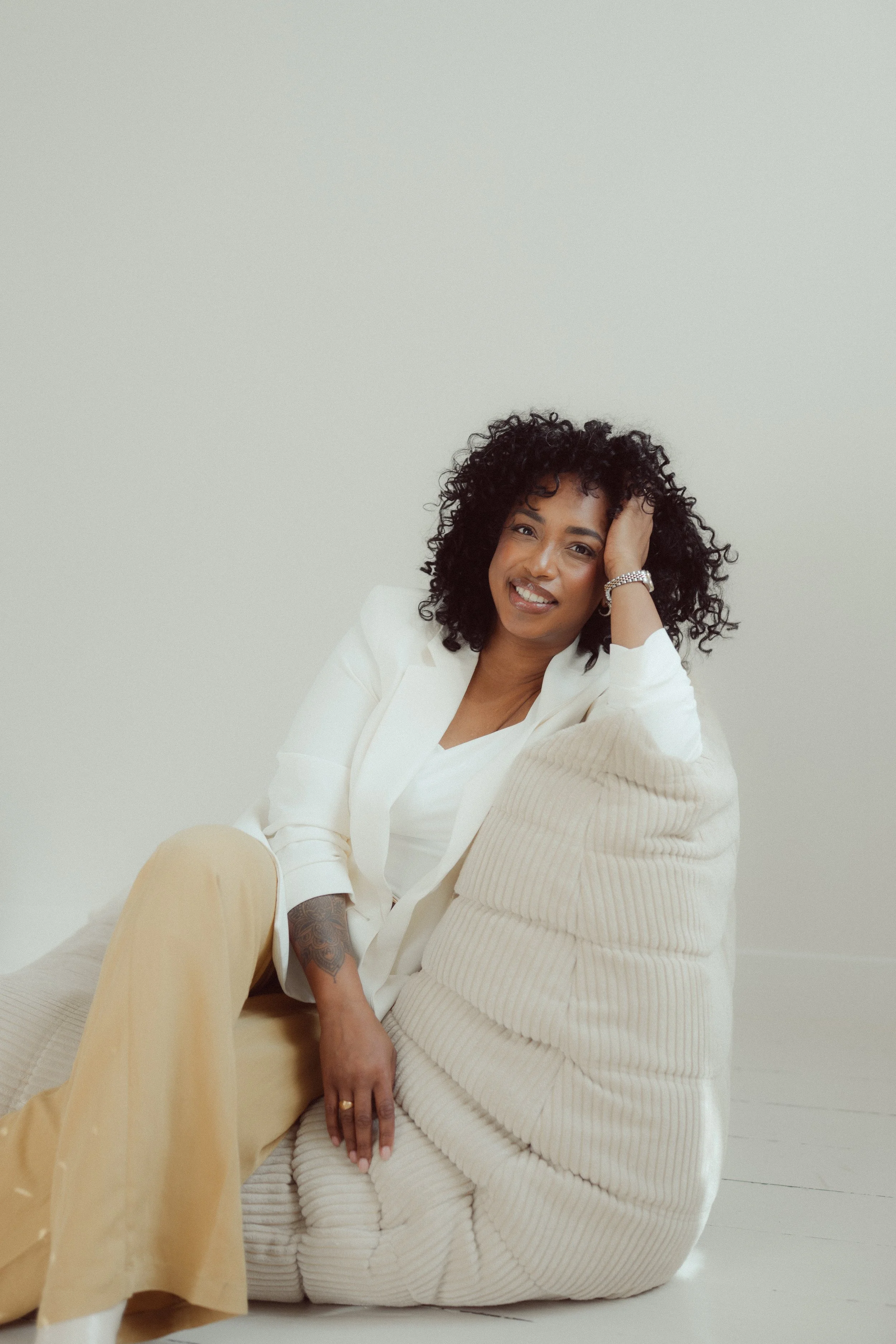 Een vrouw met krullend zwart haar, gekleed in een witte blazer en beige broek, zit op de grond en leunt tegen een vrouw in een beige ribbelige jurk. Ze lachen en kijken naar de camera.