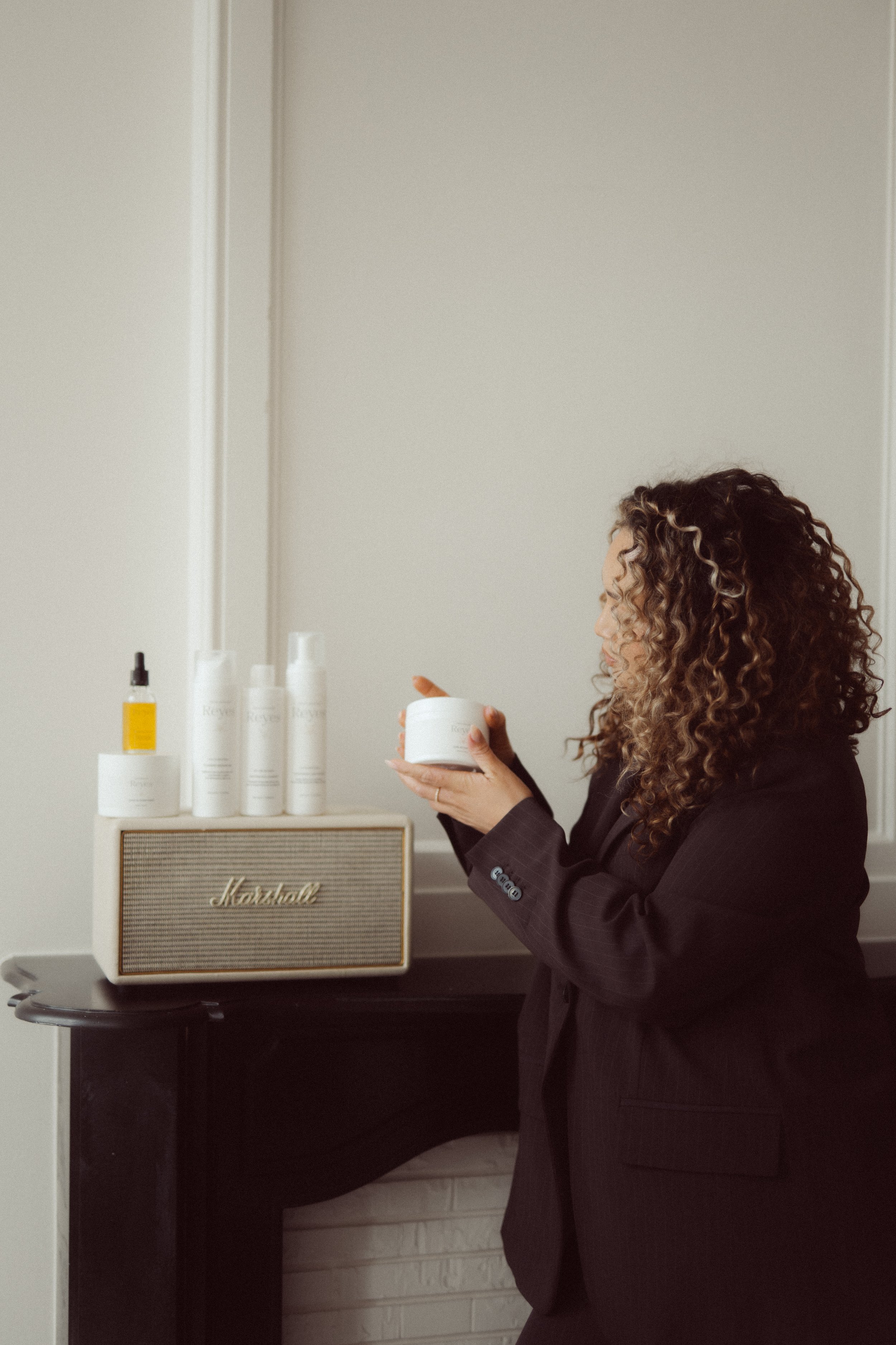 Een vrouw met krullend haar die een crèmecontainer vasthoudt in een kamer met een witte muur en een radiobadder met de merknaam Marshall.
