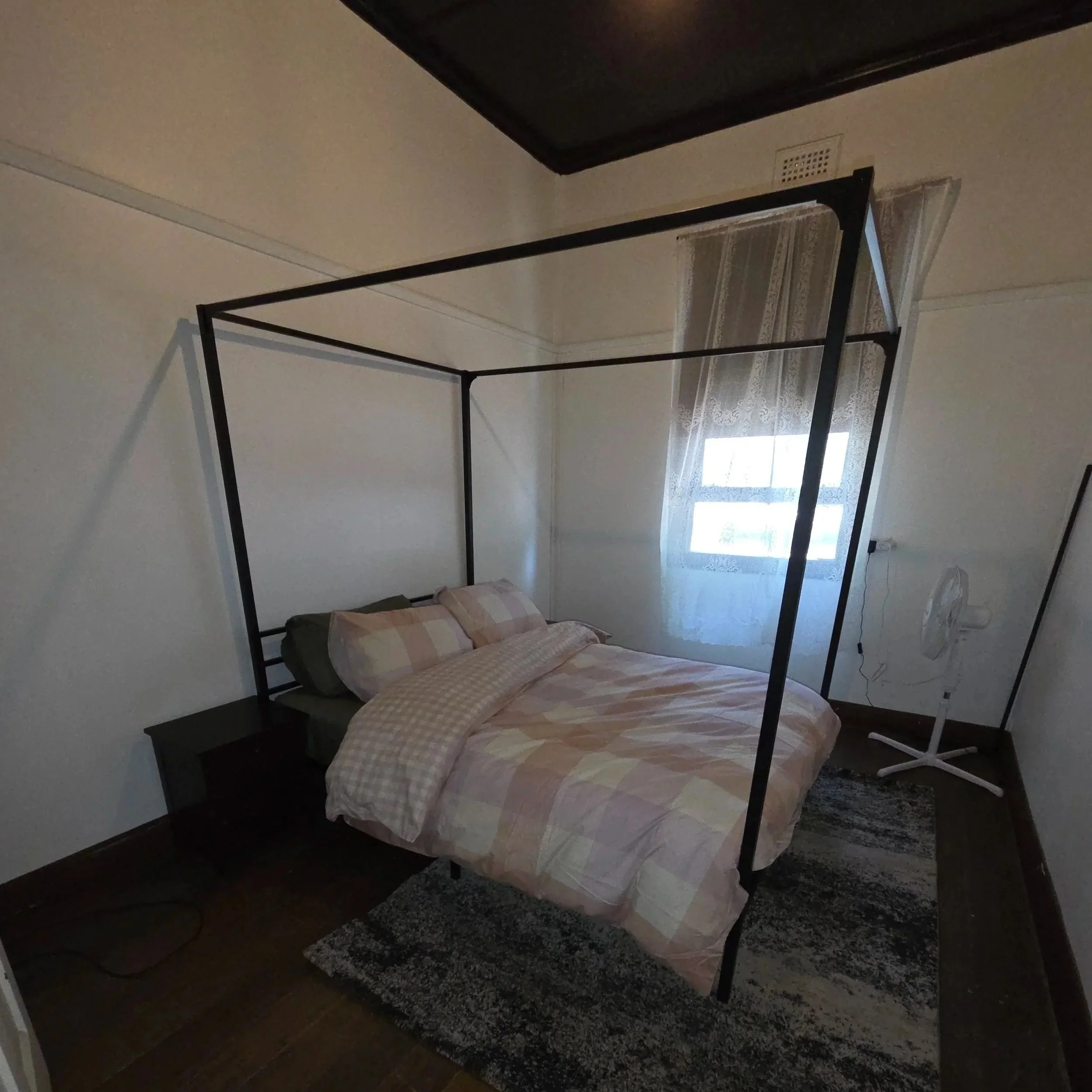 A bedroom featuring a black metal canopy bed with a pastel plaid bedspread, a small black nightstand, a window with lace curtains, a white standing fan, and a gray rug on wooden flooring.
