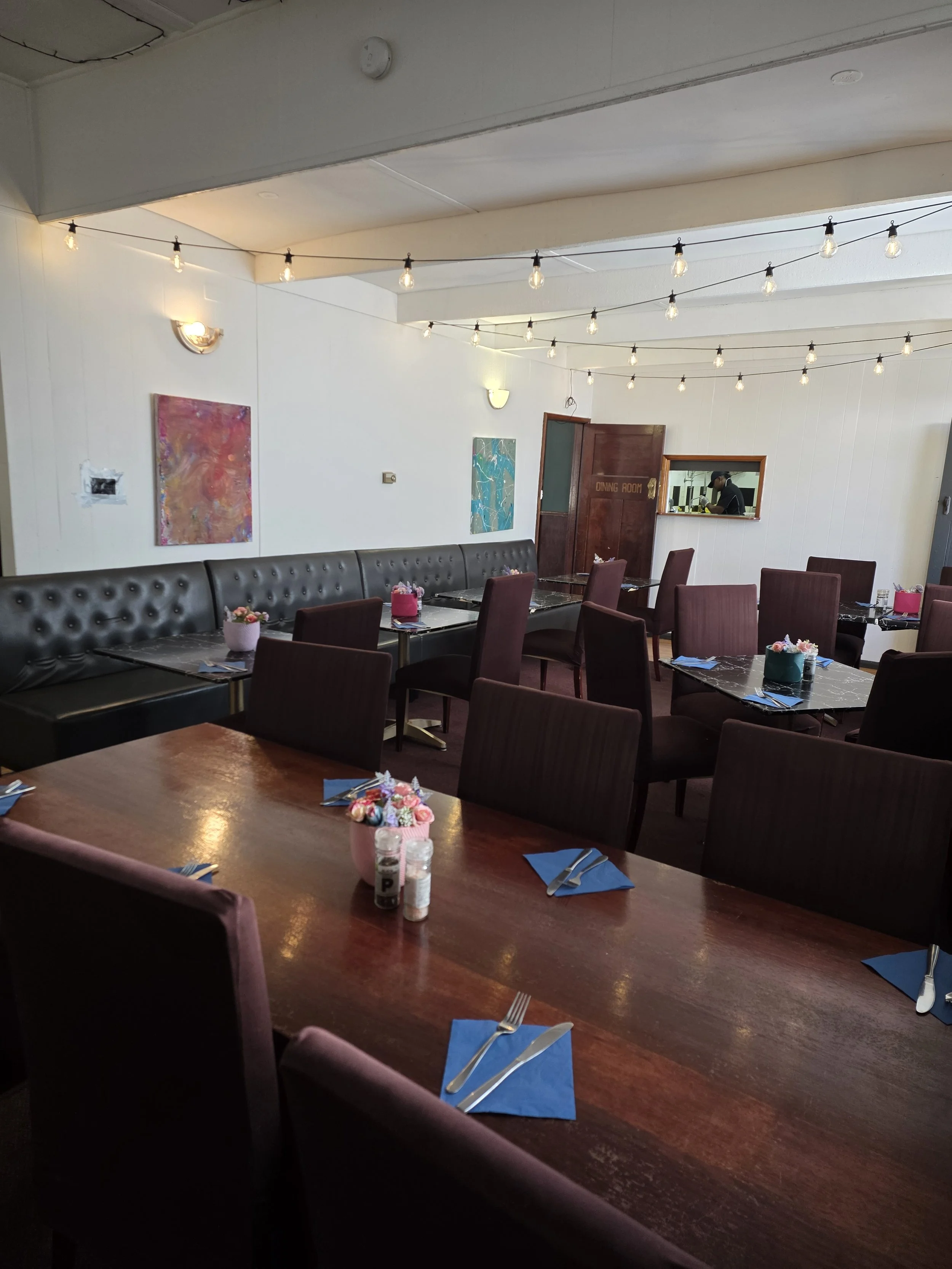Empty restaurant with pink, teal, and black chairs, black marble-topped tables, blue napkins, and pastel flower centerpieces, decorated with string lights and abstract art on white walls.