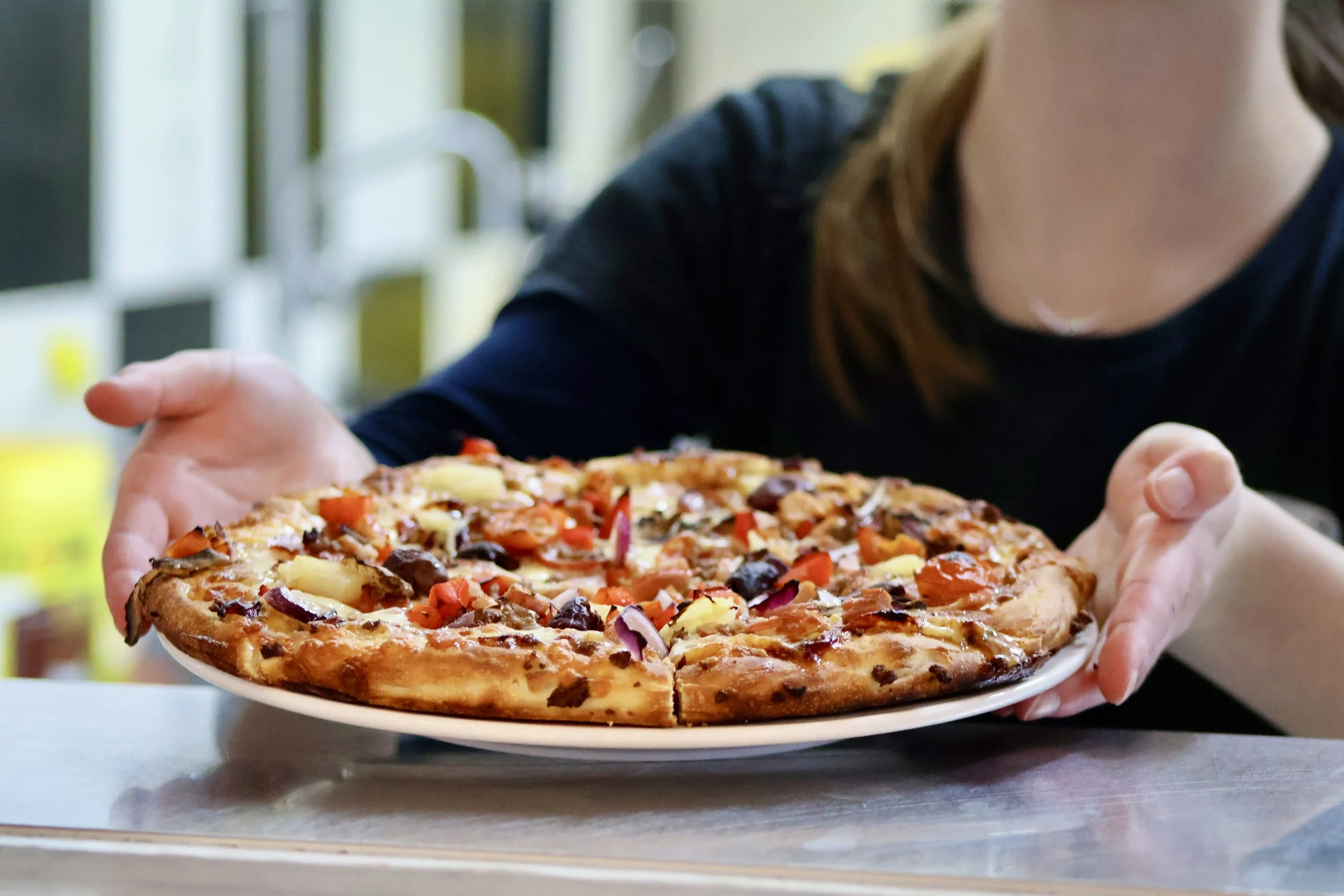 Person holding a freshly baked pizza topped with cheese, peppers, onions, mushrooms, and olives.
