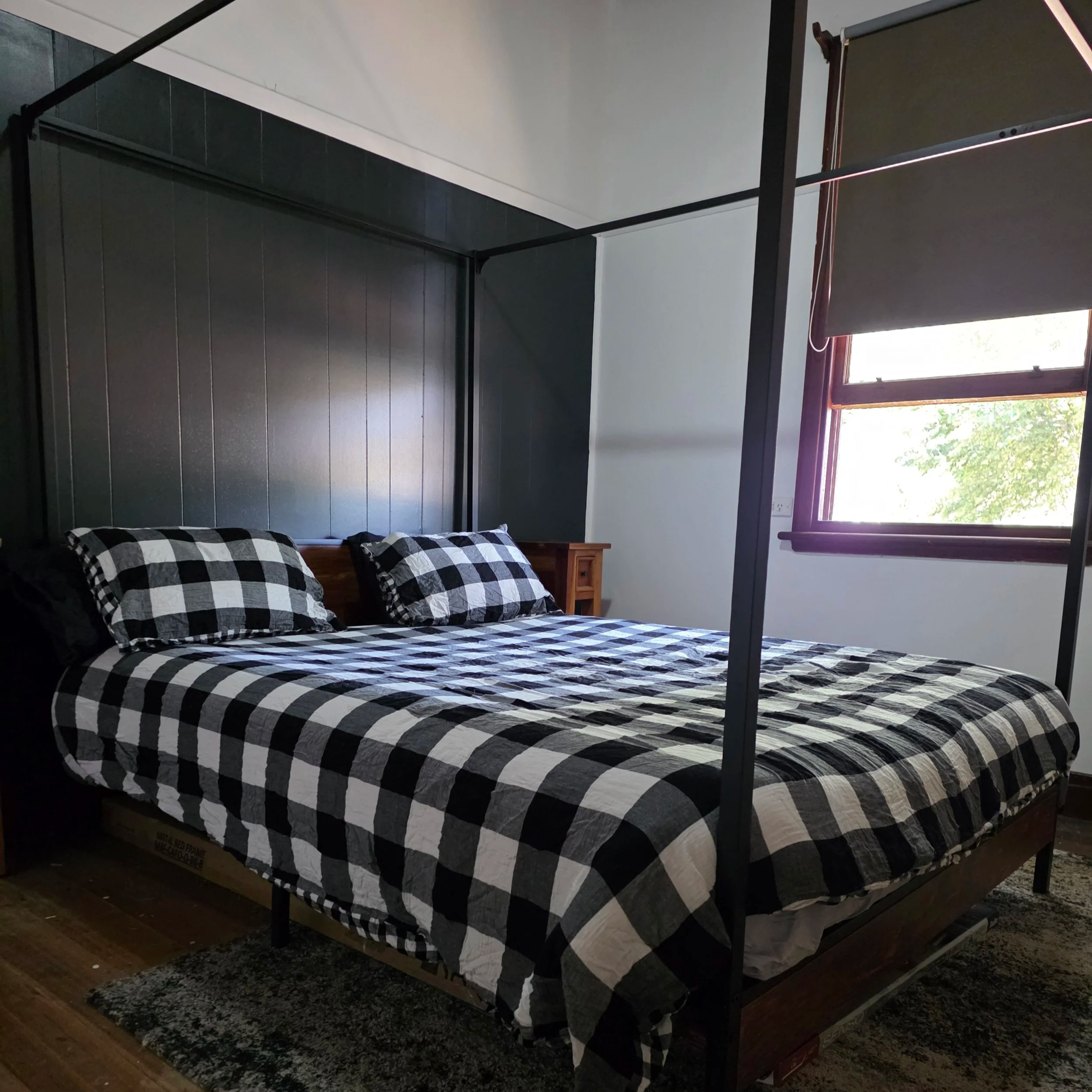 A bedroom with a black metal four-poster bed, black and white checkered bedding, and checkered pillows. There is a window with a black shade letting in natural light, and a small wooden nightstand beside the bed.