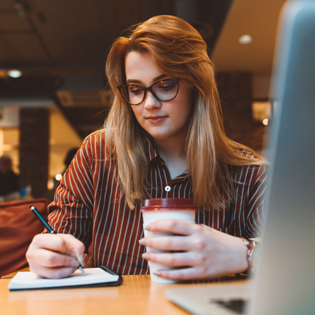 Woman wearing a striped shirt and wearing glasses in a coffee shop with coffee writing in a note pad. Stripe Social Pinterest Strategy