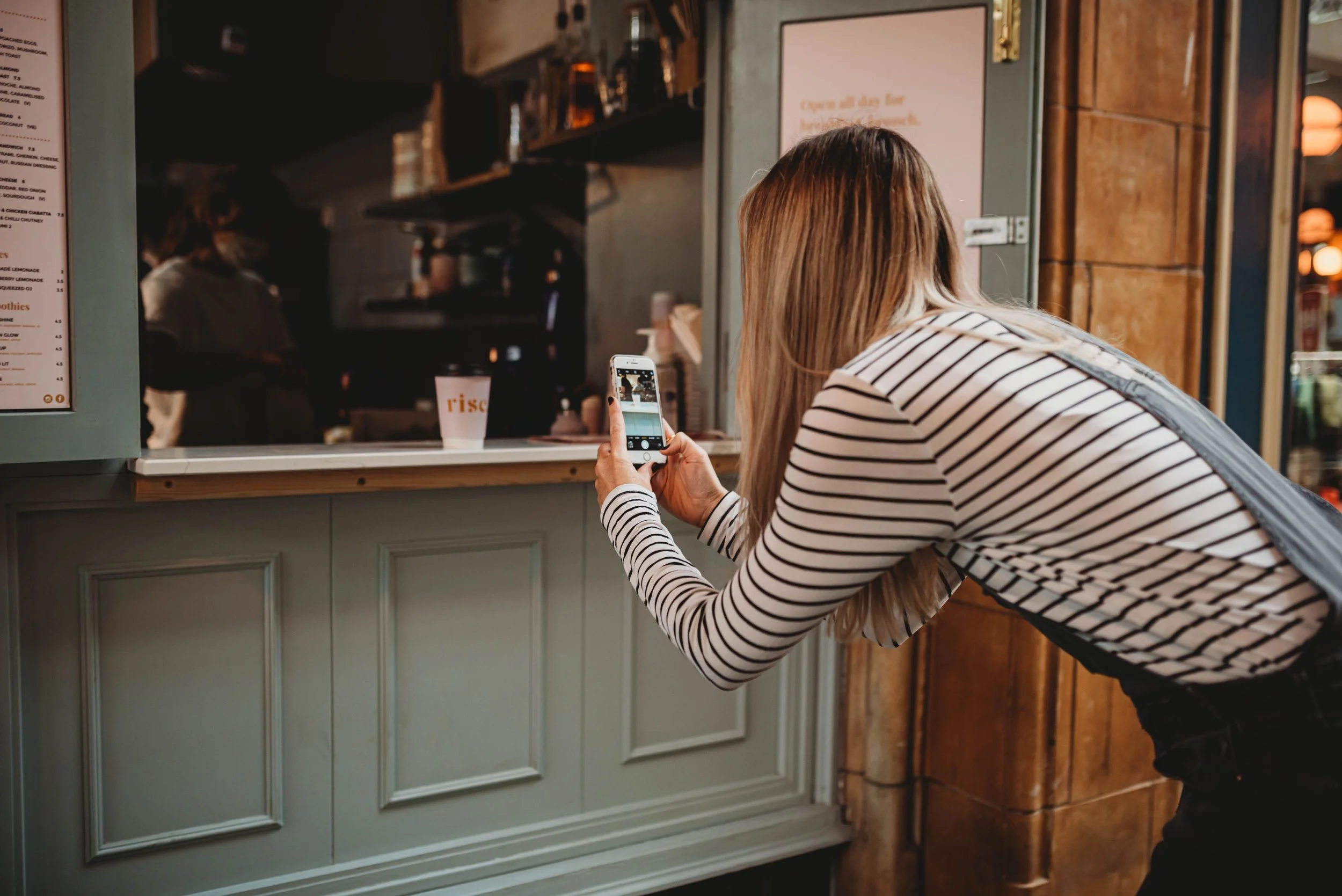 Kat from stripe social facing away from the camera wearing a striped top. She's taking a photo with her mobile phone of a take out coffee on the side of the cafe.