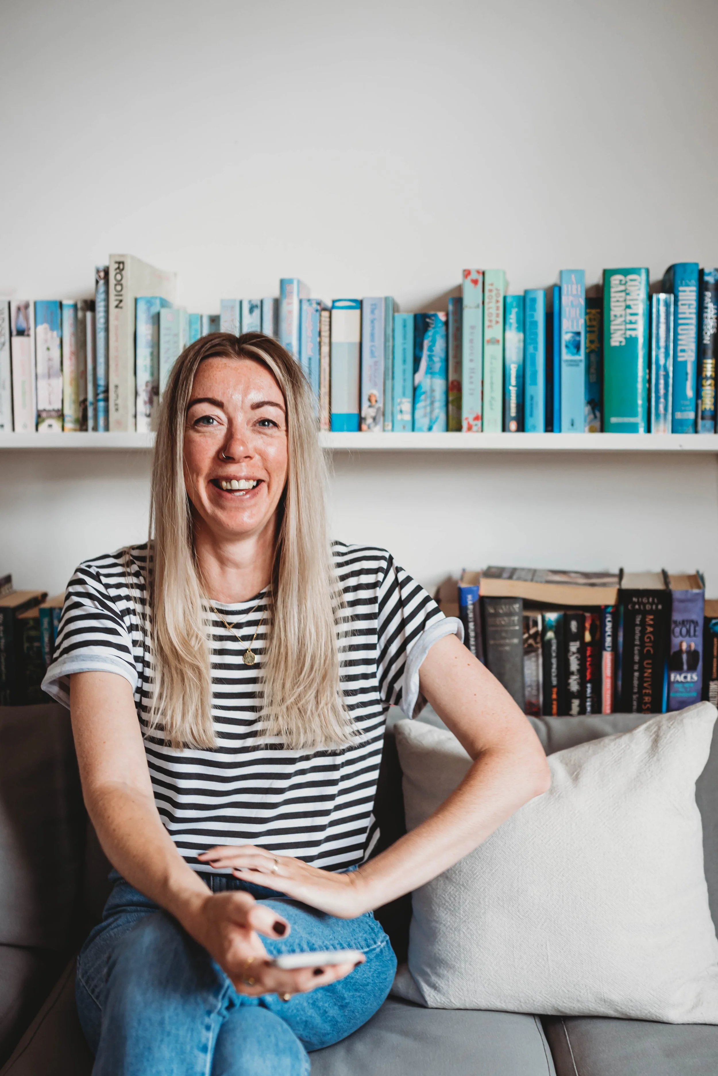 Kat from Stripe Social in a striped tshirt, holding a phone, smiling at the camera in a cafe with books in the background.