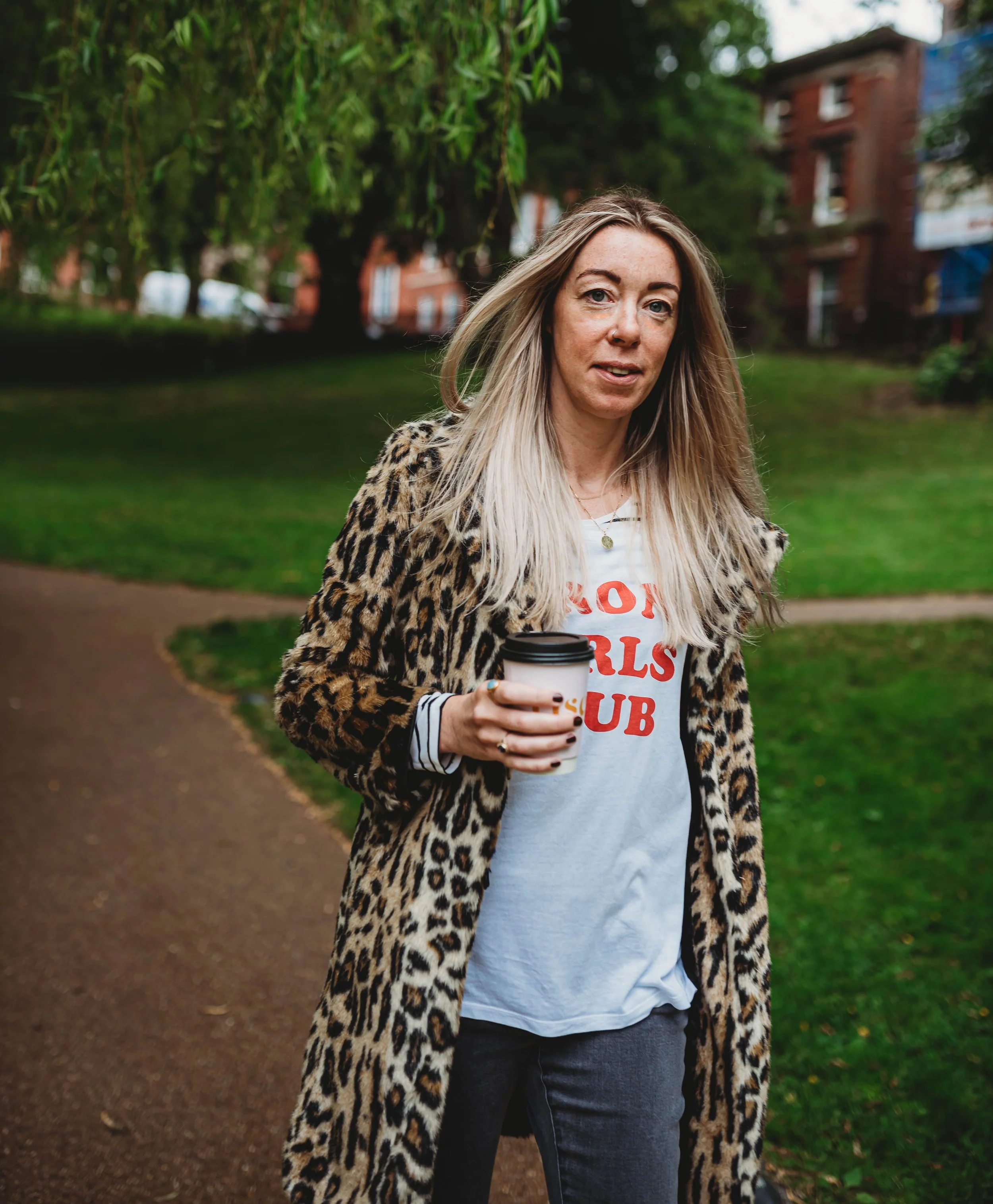 Kat, Instagram and Pinterest marketing expert, walking through a park holding a takeaway coffee wearing a leopard print coat and strong girls club t-shirt