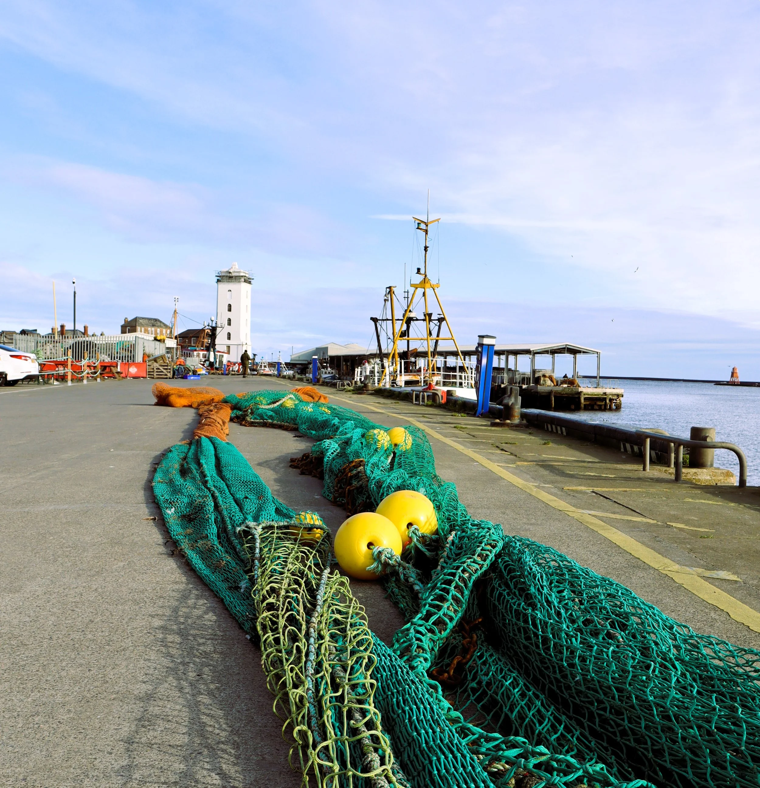 North Shields fish quay with green fishing nets and yellow buoys laid out to dry with the fishing boats in the quay behind and a white tower which is the old low light heritage centre