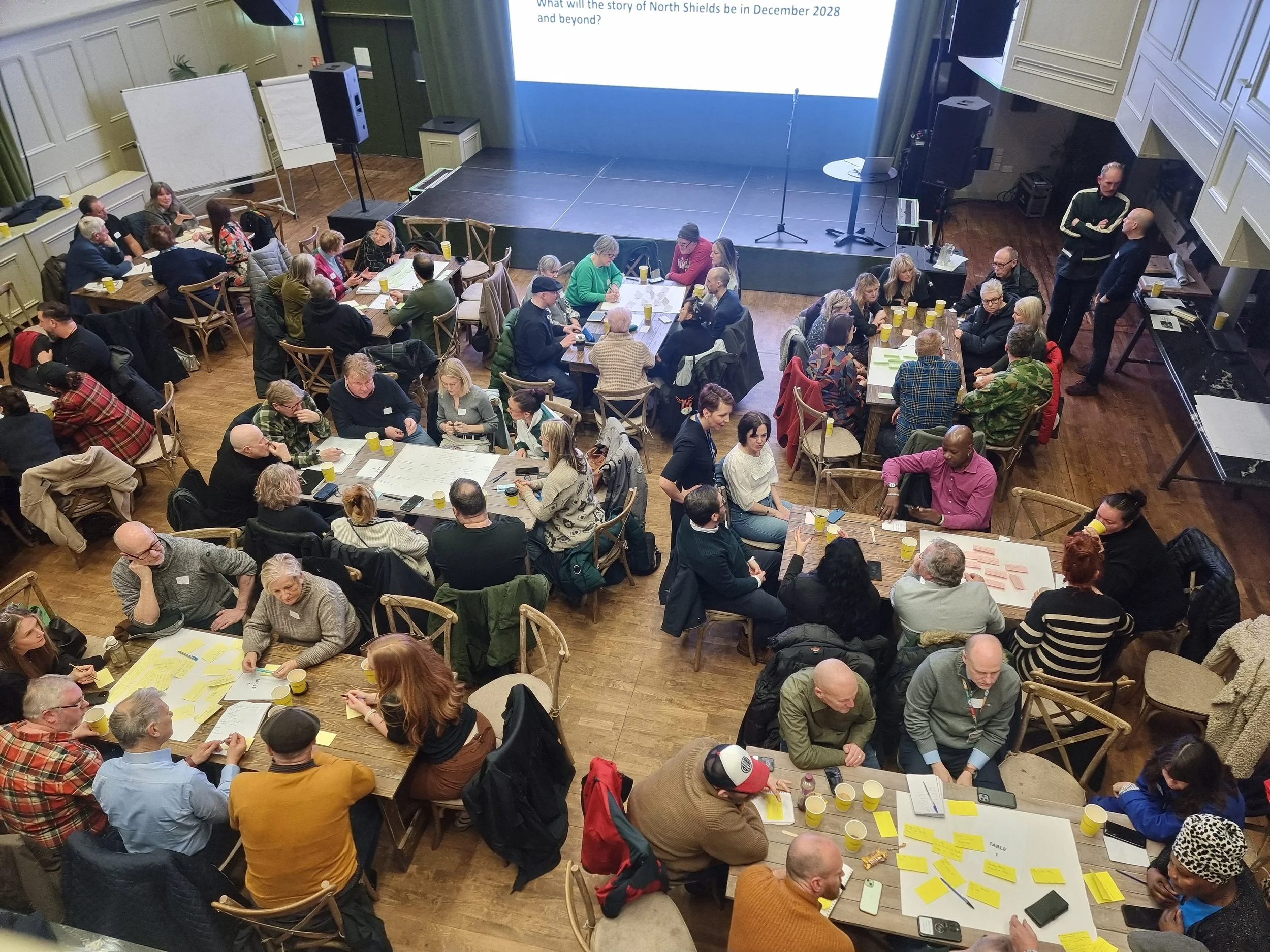 A photo taken from above showing the auditorium at the Exchange Theatre filled with tables and people of all ages sat around discussing and writing on A1 sheets andfpost it notes