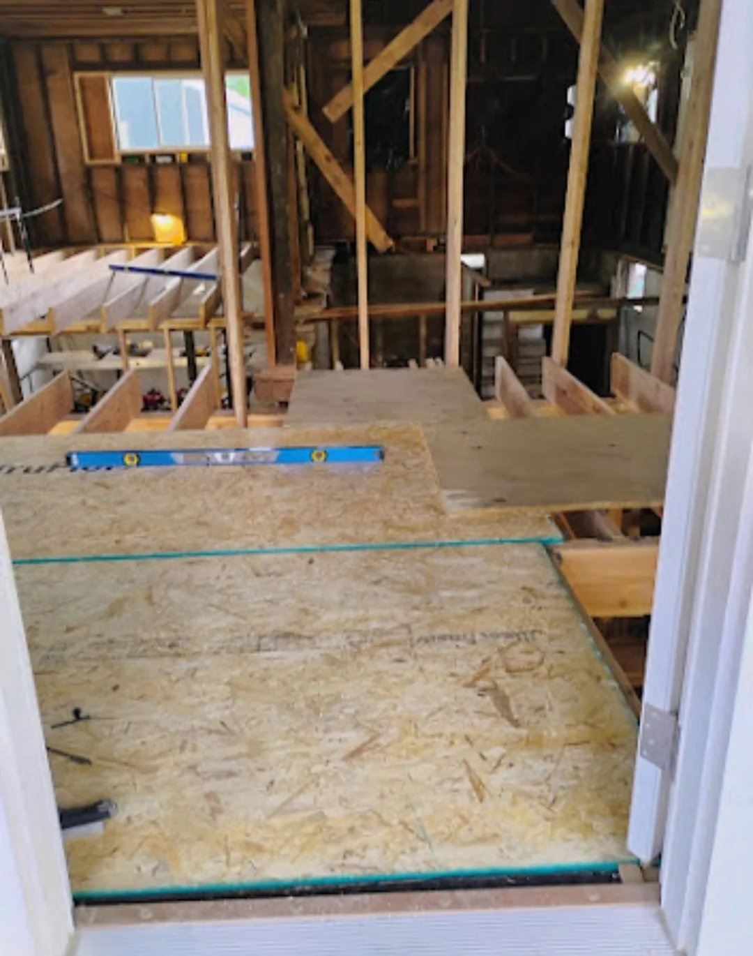 Inside a house under construction, showing exposed wooden framing and subflooring.