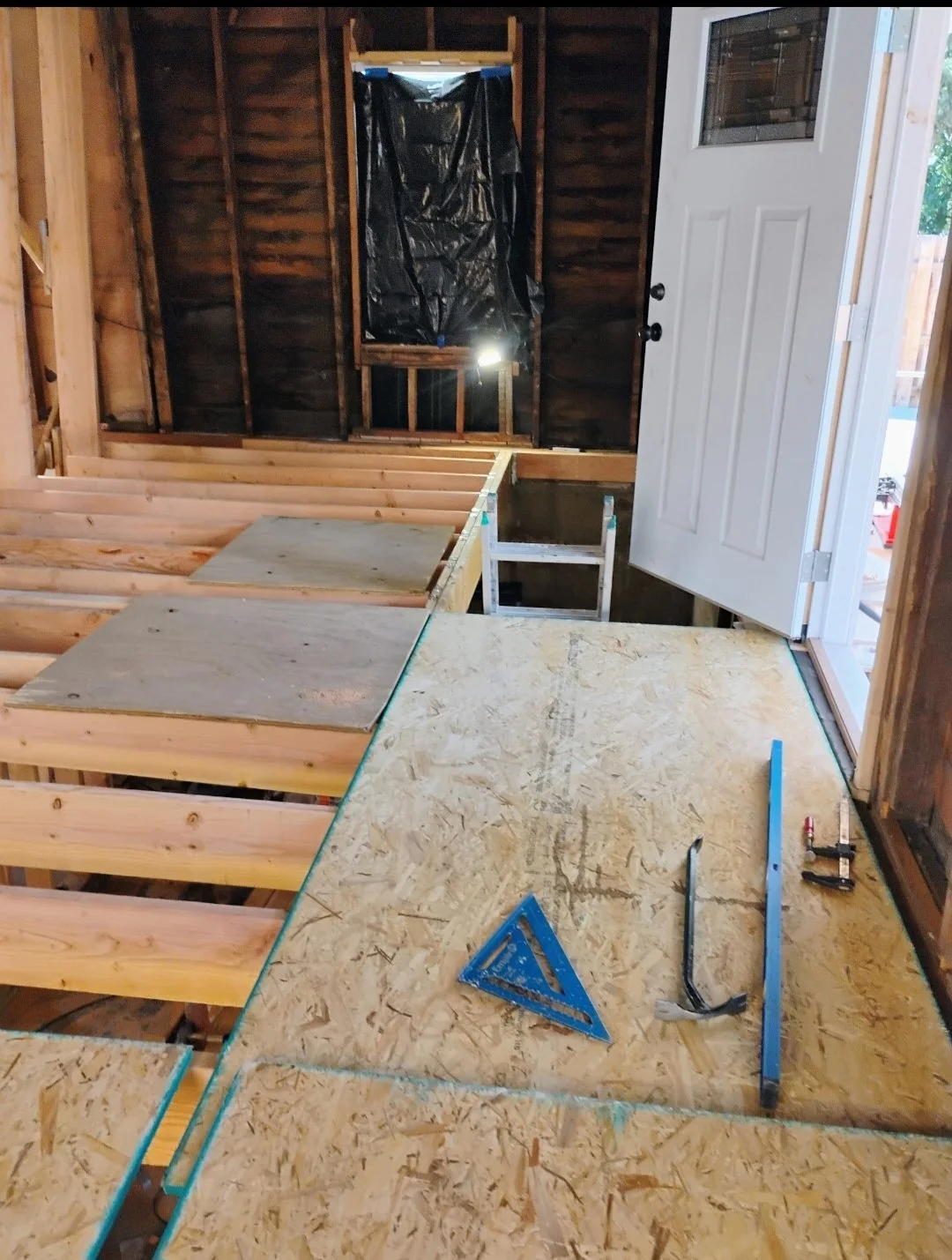 Interior of a house under construction with partially built floor, tools, and an open door leading outside.