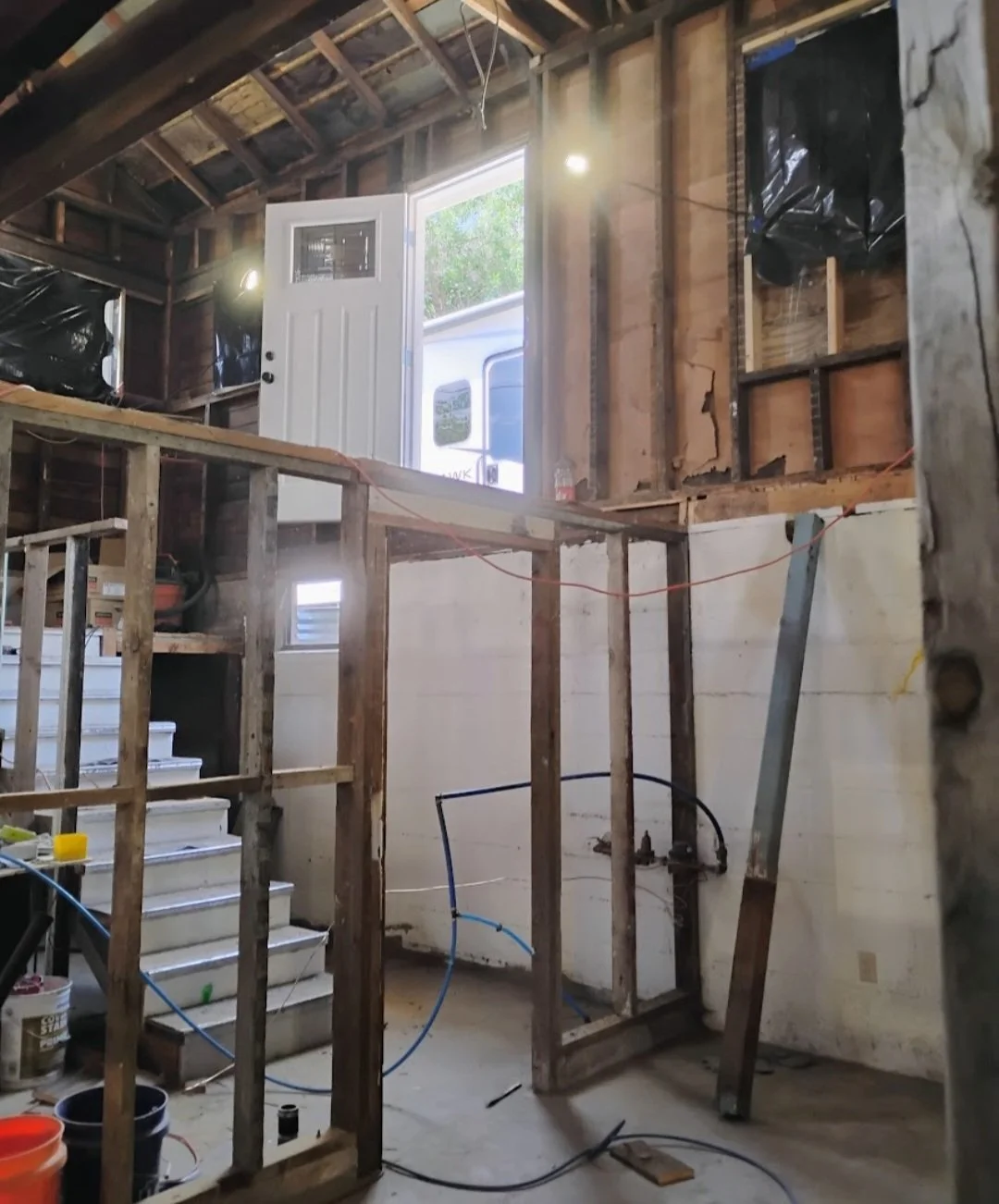 Interior of a house under renovation or construction, showing exposed wooden framing, a staircase, and a door open to outside with sunlight.