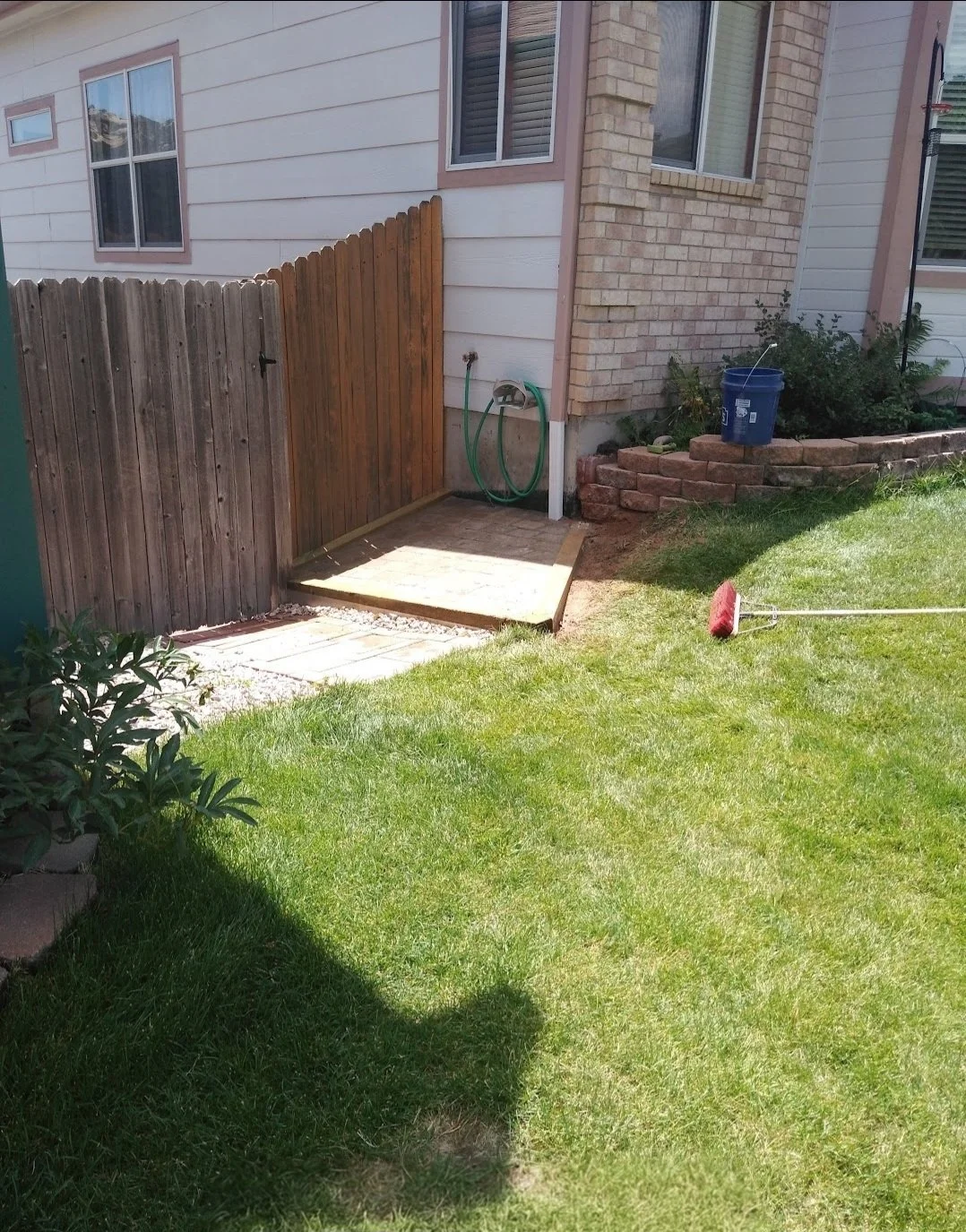 Backyard with newly built small wooden deck area, green garden hose hanging on house wall, blue bucket on flower bed, and red broom laying on grass.