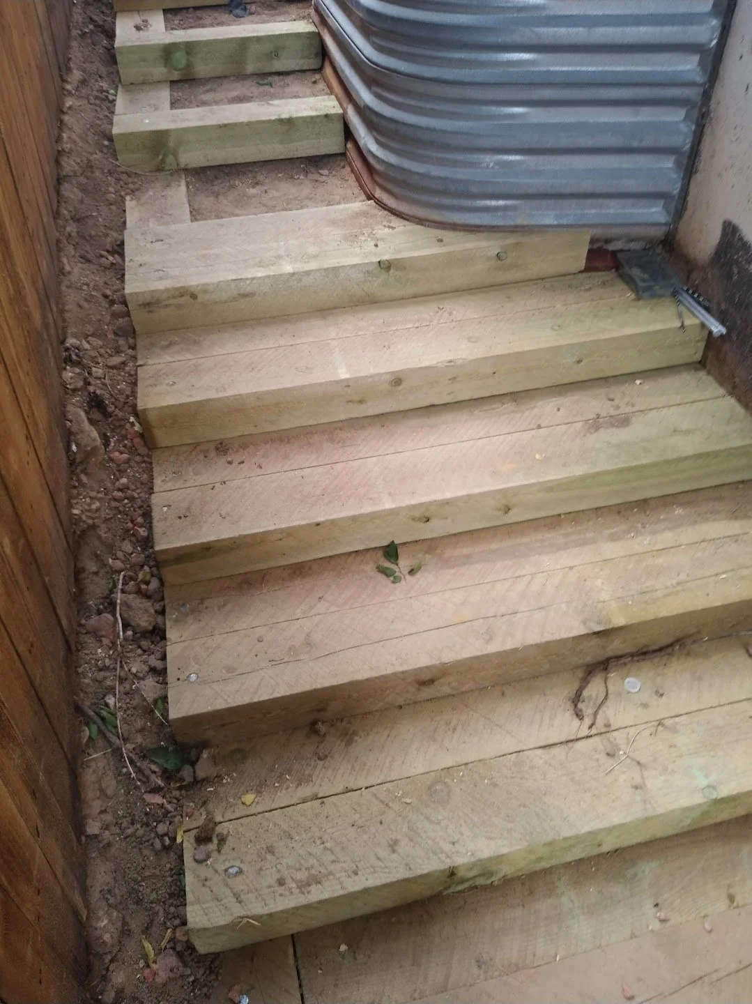 Wooden steps being installed outdoors, partially constructed with visible screws, next to a corrugated metal wall and a wooden fence.