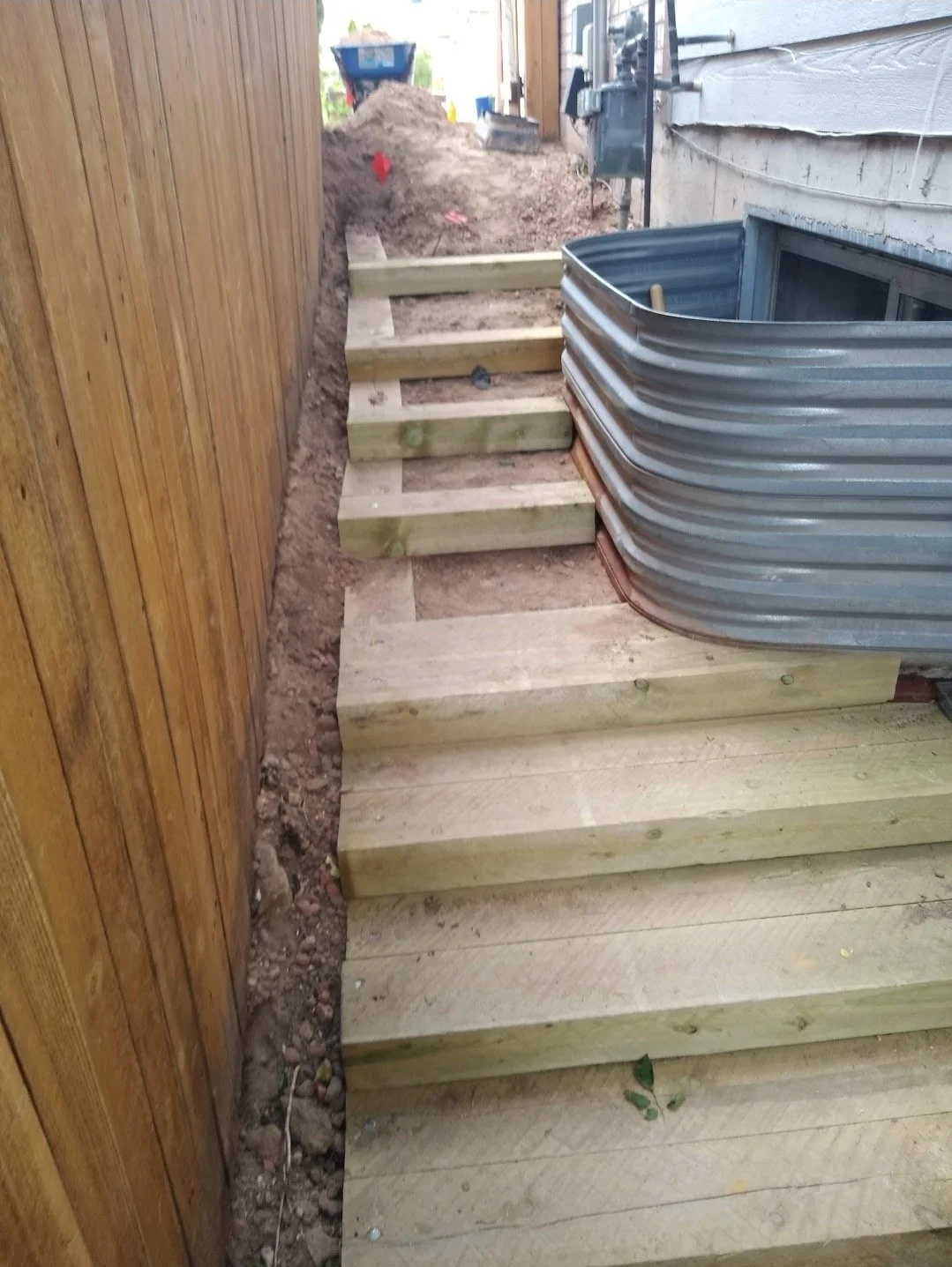 Wooden steps under construction next to an exterior house wall, with a metal corrugated pipe nearby and construction materials in the background.