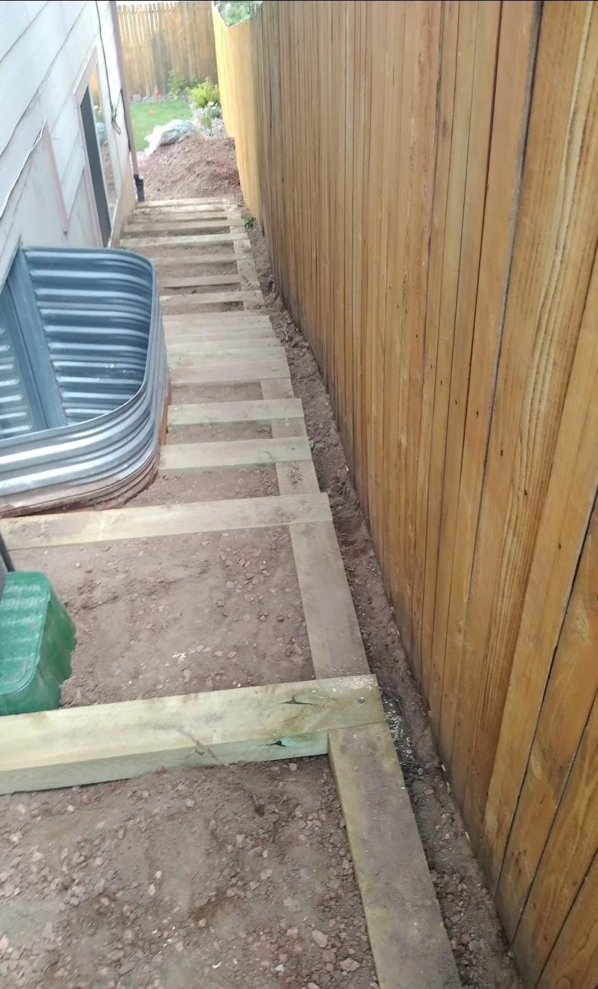 Side view of a narrow outdoor walkway under construction with wooden framing and a tall wooden fence.