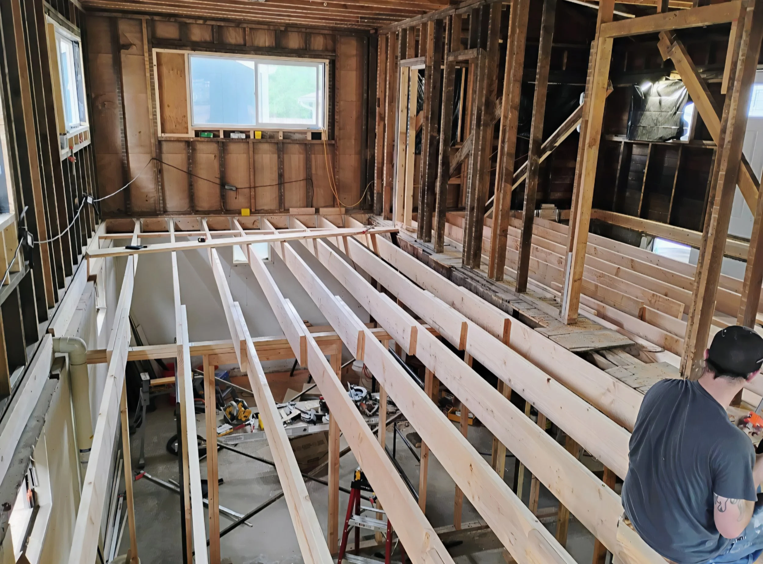 Interior of a house under construction with exposed wooden framing and a partially installed staircase. Male worker in helmet is visible on lower right corner.