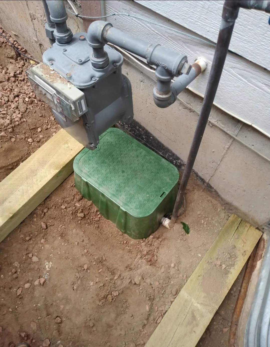 Outdoor utility meter box with piping, mounted on a wall, next to a green cable cover and wooden framing, with soil and a small green leaf on the ground.