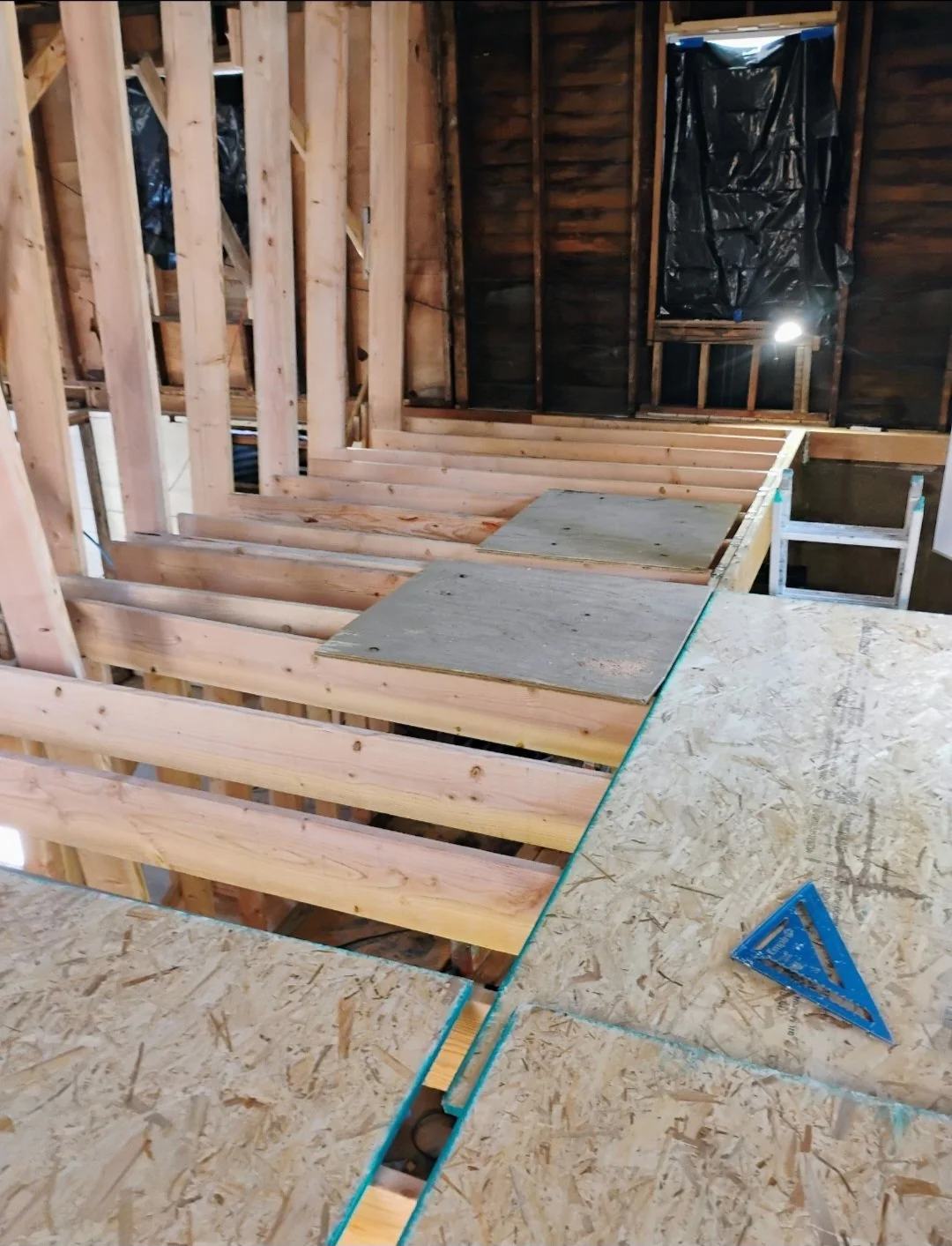 Interior view of a house under construction showing wooden floor framing and drywall plywood sheets. Framing walls and a window opening are visible, with black plastic vapor barrier on the wall.