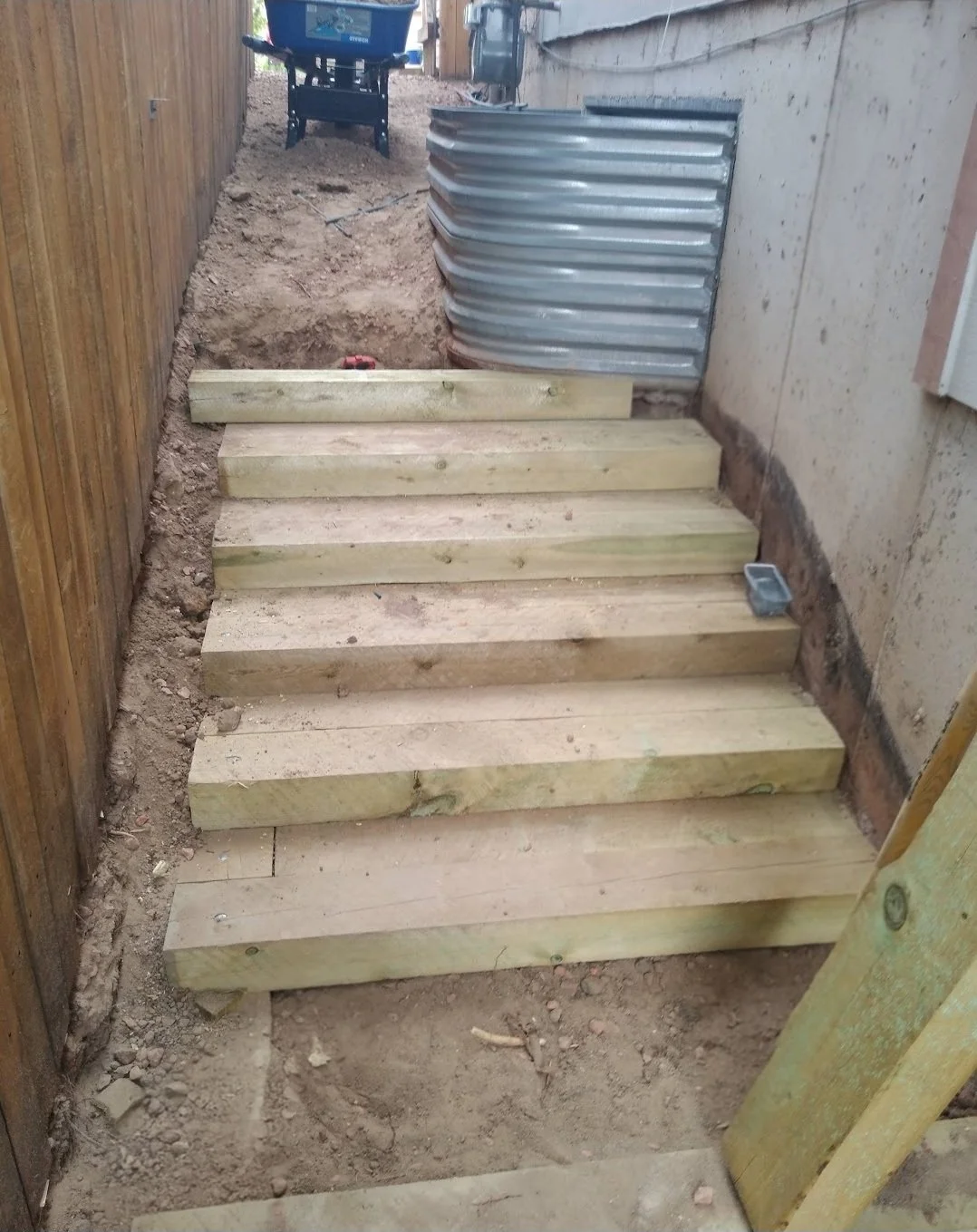 Wooden staircase under construction with several wooden planks laid on the ground for steps, a metal sheet barrier to the side, and construction tools nearby.