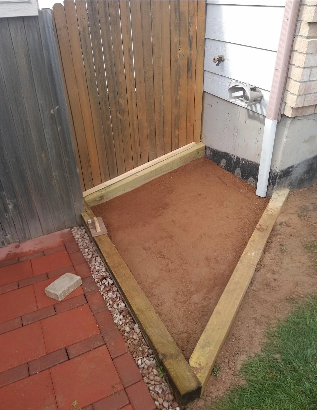 A partially constructed outdoor shower stall with a wooden frame, sand base, nearby brick and grass, and a brick wall and wooden fence in the background.
