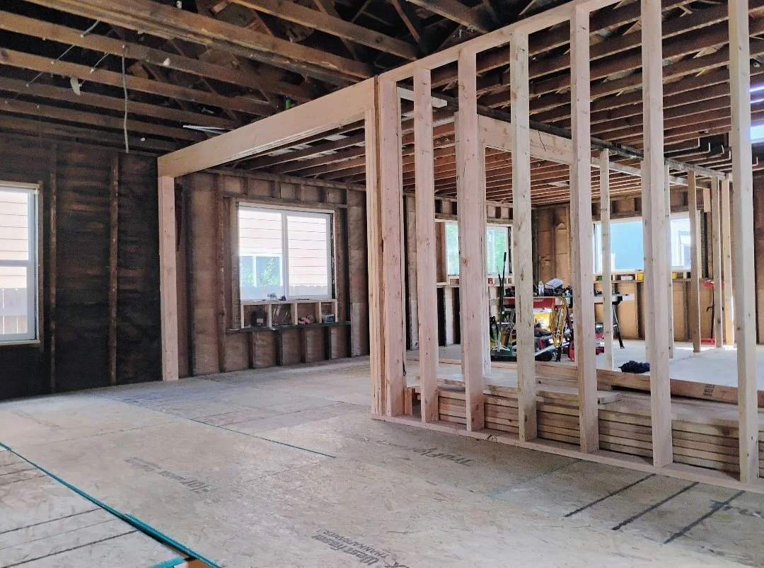Interior of a house under construction, showing exposed framing, plywood subflooring, and two windows.