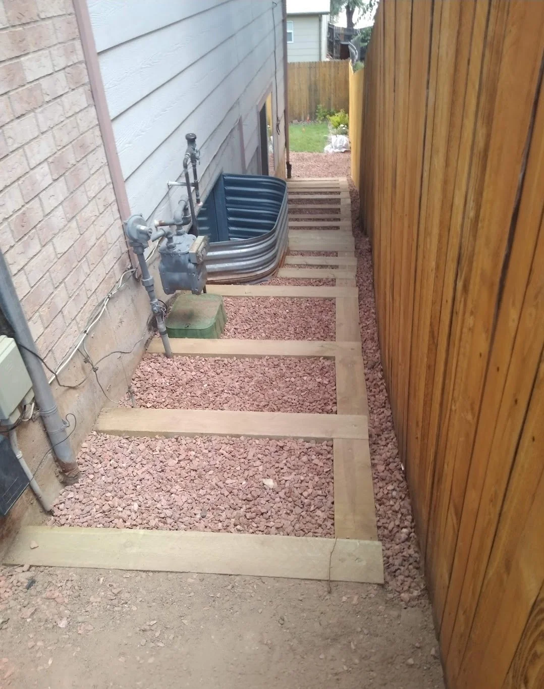 Side yard with a pathway under construction, wooden frame borders, pink gravel, blue plastic basket, and part of the house's exterior wall.