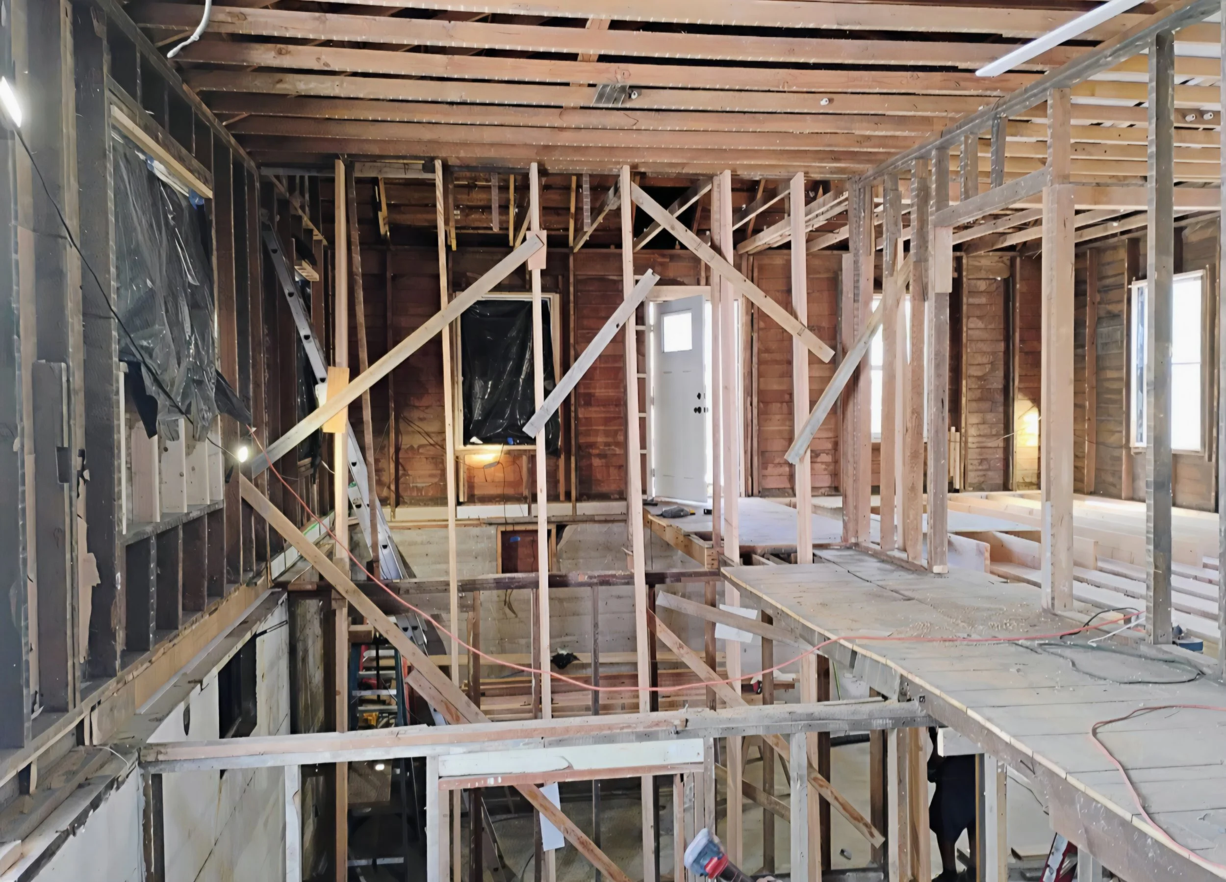 Interior view of a house under construction with exposed wooden framing, studs, and ceiling beams, and some wiring visible.