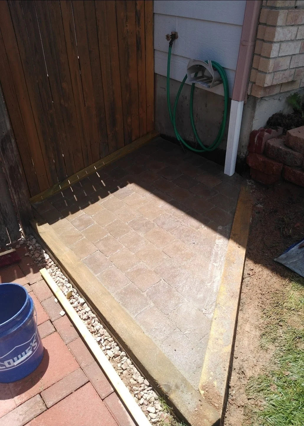 A small outdoor patio area with a brick floor, enclosed by wooden and brick walls, with a garden hose on a holder attached to a house wall.