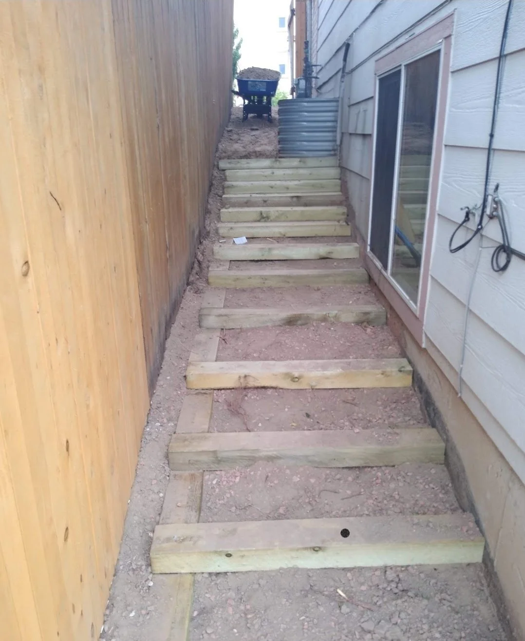Wooden steps under construction between a sandstone house and a wooden fence, with dirt ground and a wheelbarrow at the top.