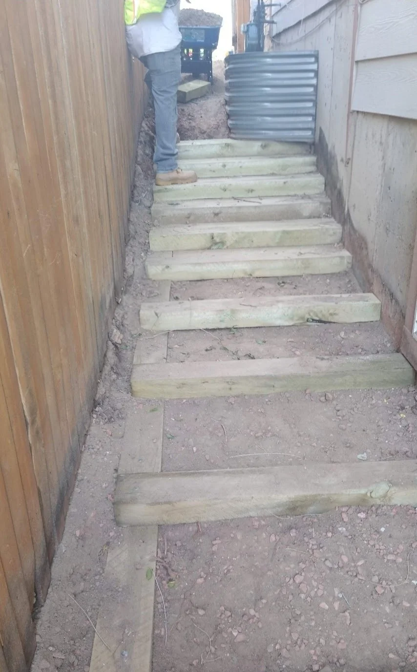 Construction site showing a narrow outdoor staircase made of newly installed, unfinished wooden steps between a wood fence and a house, with a person working at the top of the stairs.