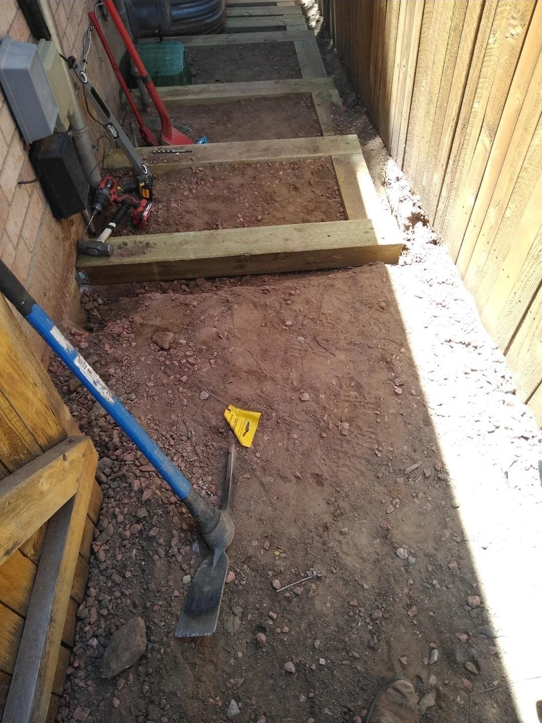 A small garden or walkway area with wooden frames on the ground, construction tools including a shovel and power drill, and a partially enclosed space with a wooden fence and brick wall.