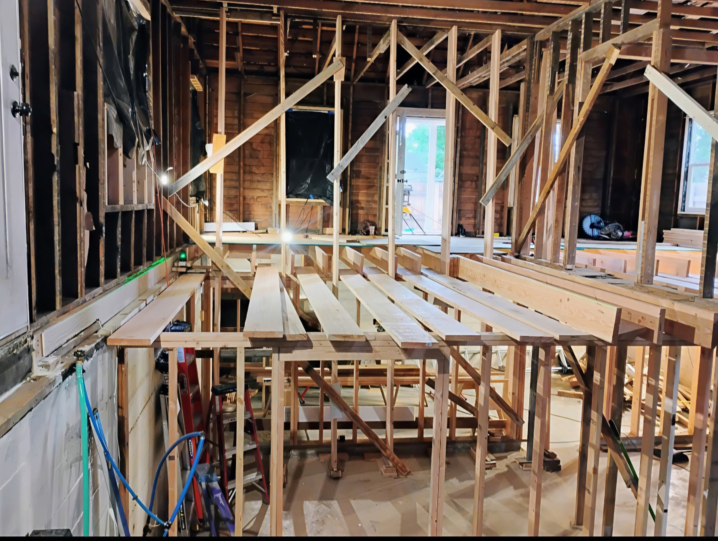 The image shows the framing structure of a house under construction, with exposed wooden beams, studs, and partial walls. Construction tools and materials are visible around the space, which has windows and an open door leading outside.