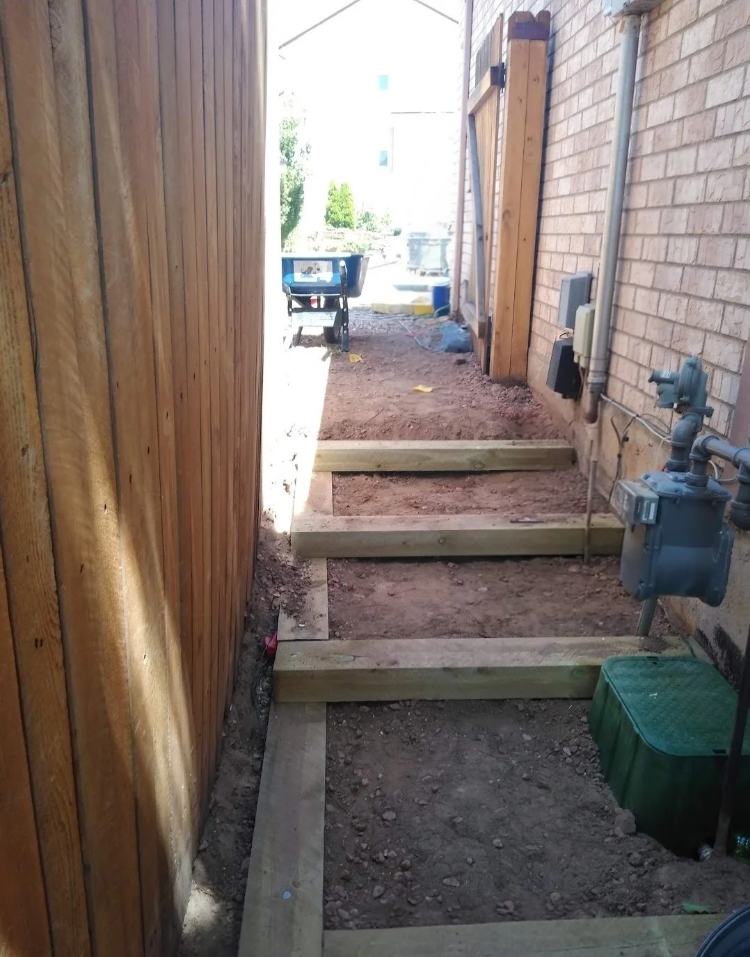 Construction site with wooden framing for a walkway or steps between a brick wall and wooden fence. Tools, materials, and utilities like a gas meter are visible.