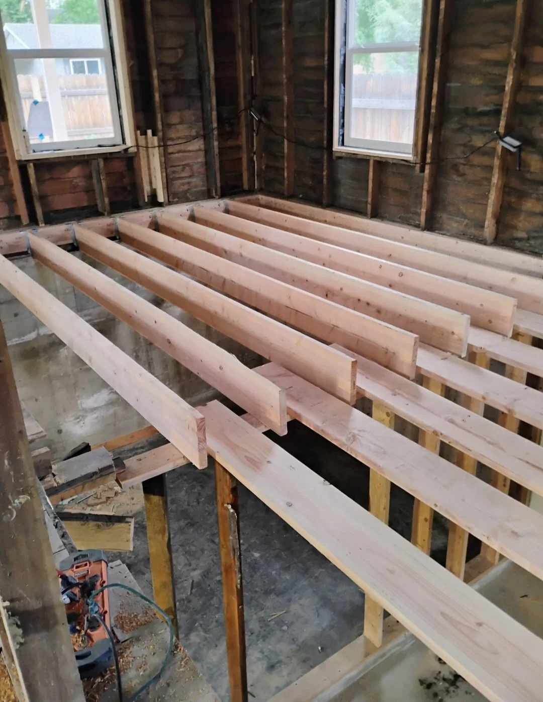 Interior of a house under renovation with wooden floor joists installed, two windows, and exposed wall framing.