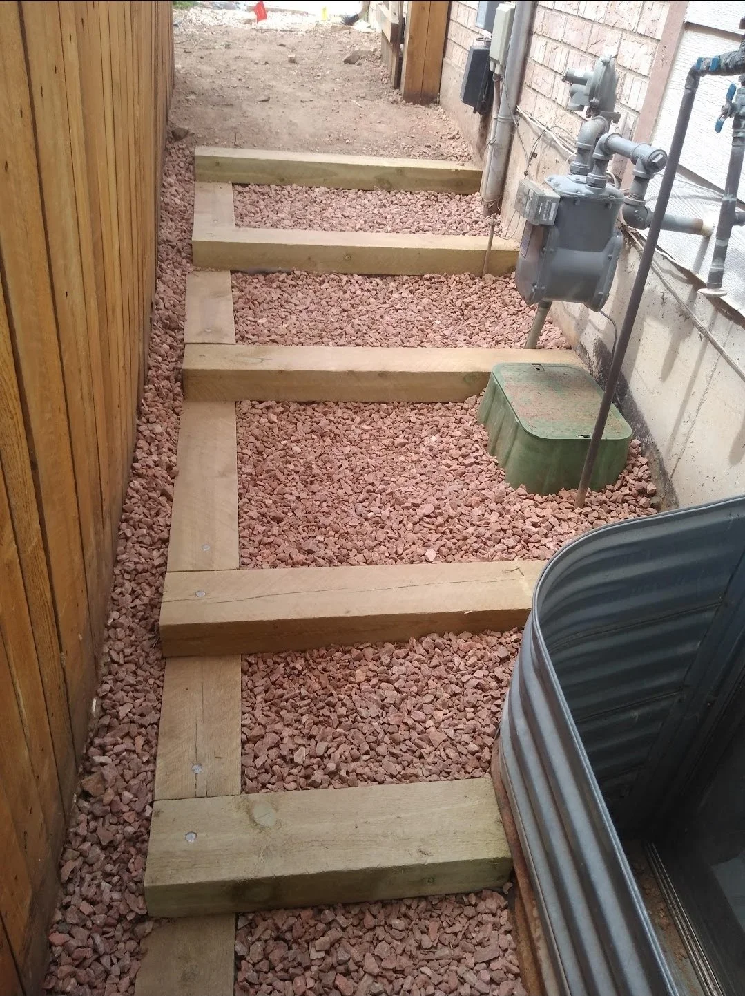 Wooden frame steps being built on reddish gravel path next to a brick and wood wall, with utility meters and pipes on the wall.