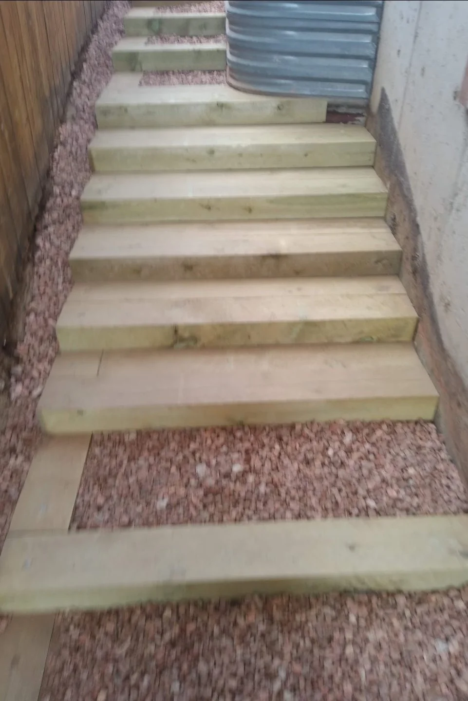 Wooden stairs under construction, with some steps installed and some remaining to be added, next to gravel ground and building walls.