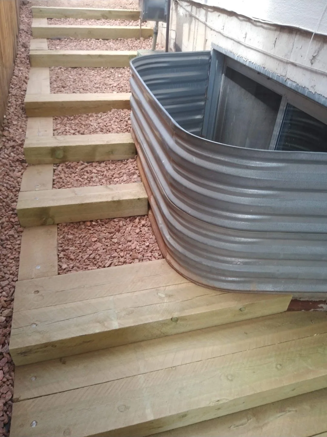 Wooden steps with gravel leading to a small metal hot tub next to a house wall.