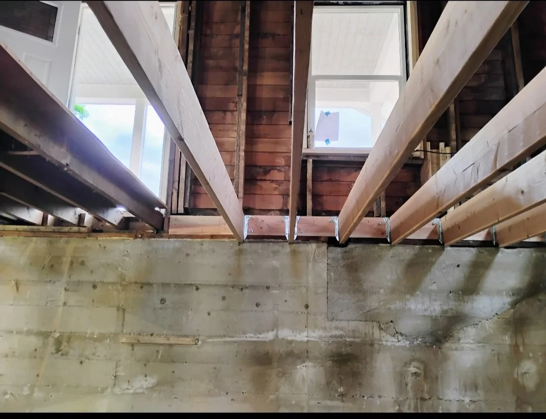 Interior of a building under construction, showing wooden framing and a concrete foundation wall, with windows and some construction materials visible.