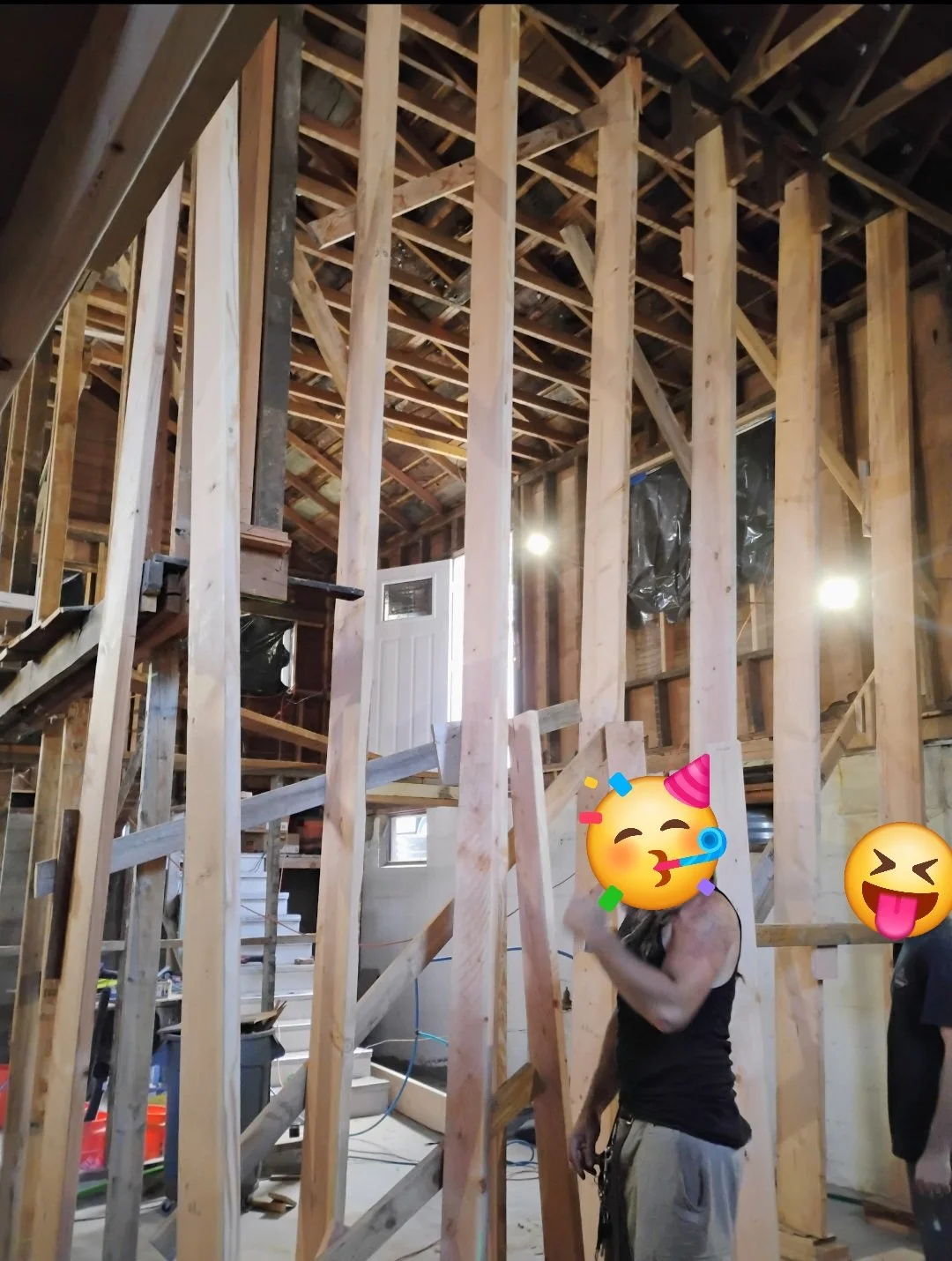Interior view of a house under construction with wooden framing, stairs, and construction workers with emojis covering their faces.