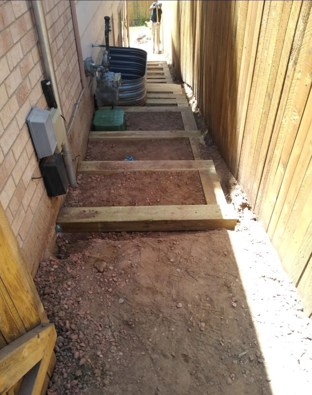 A narrow outdoor space with a dirt ground, wooden framing for a walkway or stairs, a brick wall with utility meters, and a wooden fence. There are construction materials and a blue hose reel in the background.