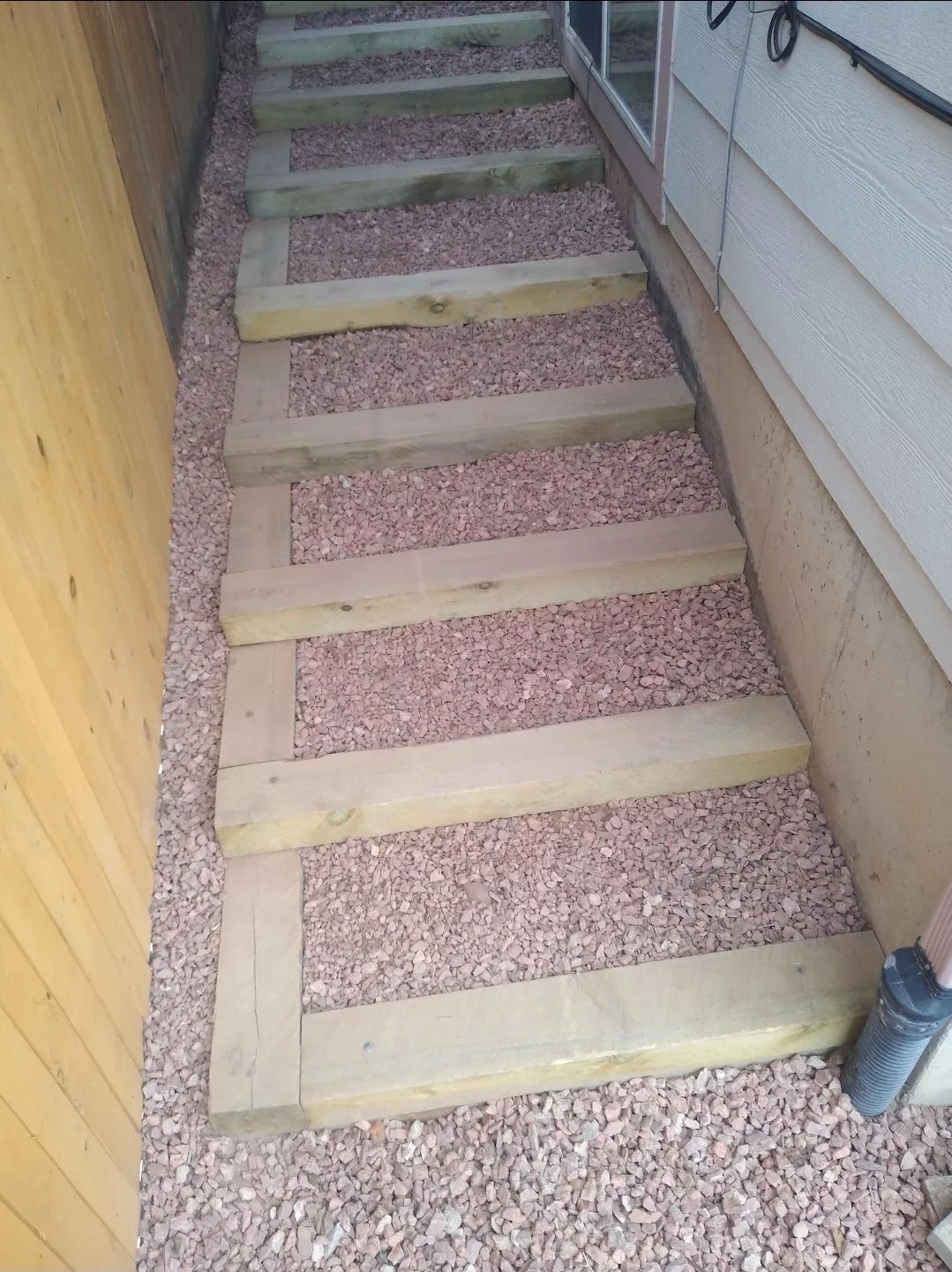 Unfinished wooden staircase frame with horizontal supports, installed on a gravel ground beside a house and next to a wooden fence.