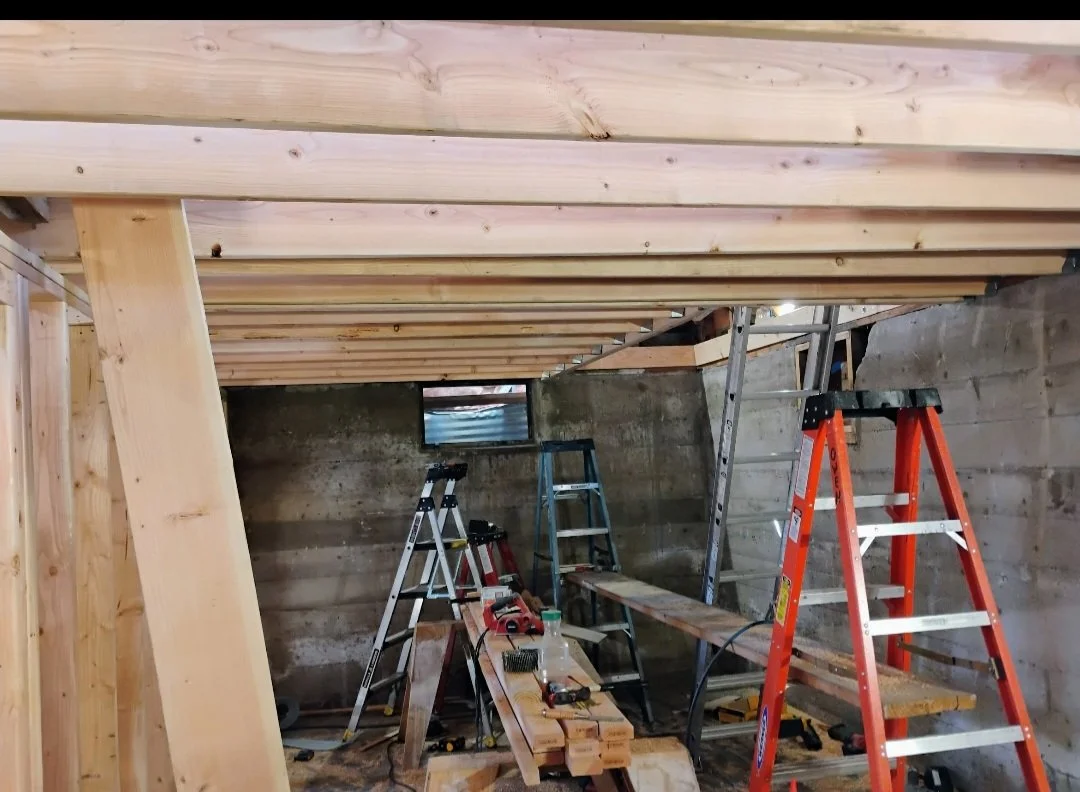 Construction site with ladders, wooden beams, tools, and a partially built wooden ceiling or loft in a basement or enclosed space.
