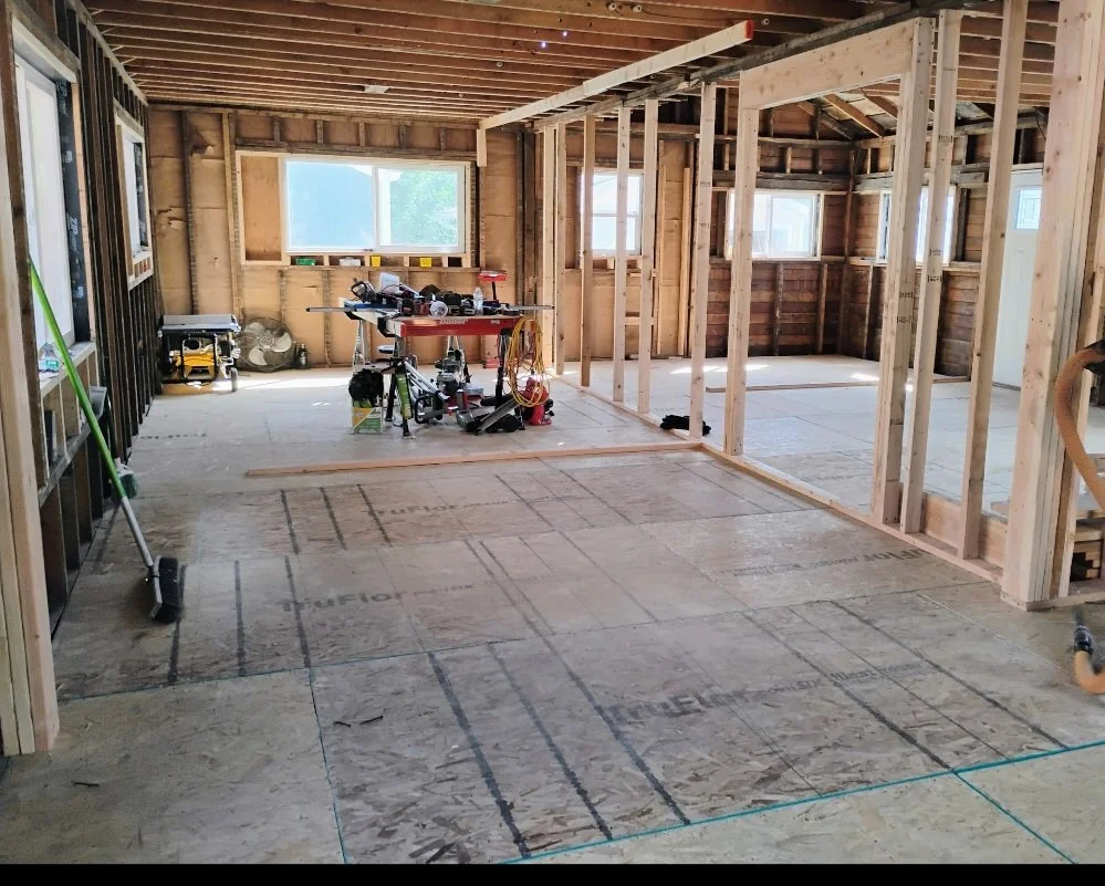 Interior of a house under construction with exposed wooden framing, construction tools on a work table, and partially installed flooring.