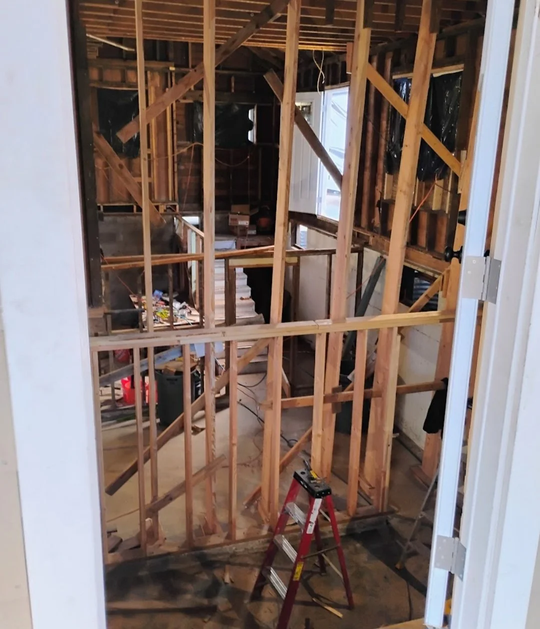 Interior of a room under construction with exposed wooden framing, a stepladder in the foreground, and construction tools and materials scattered around.