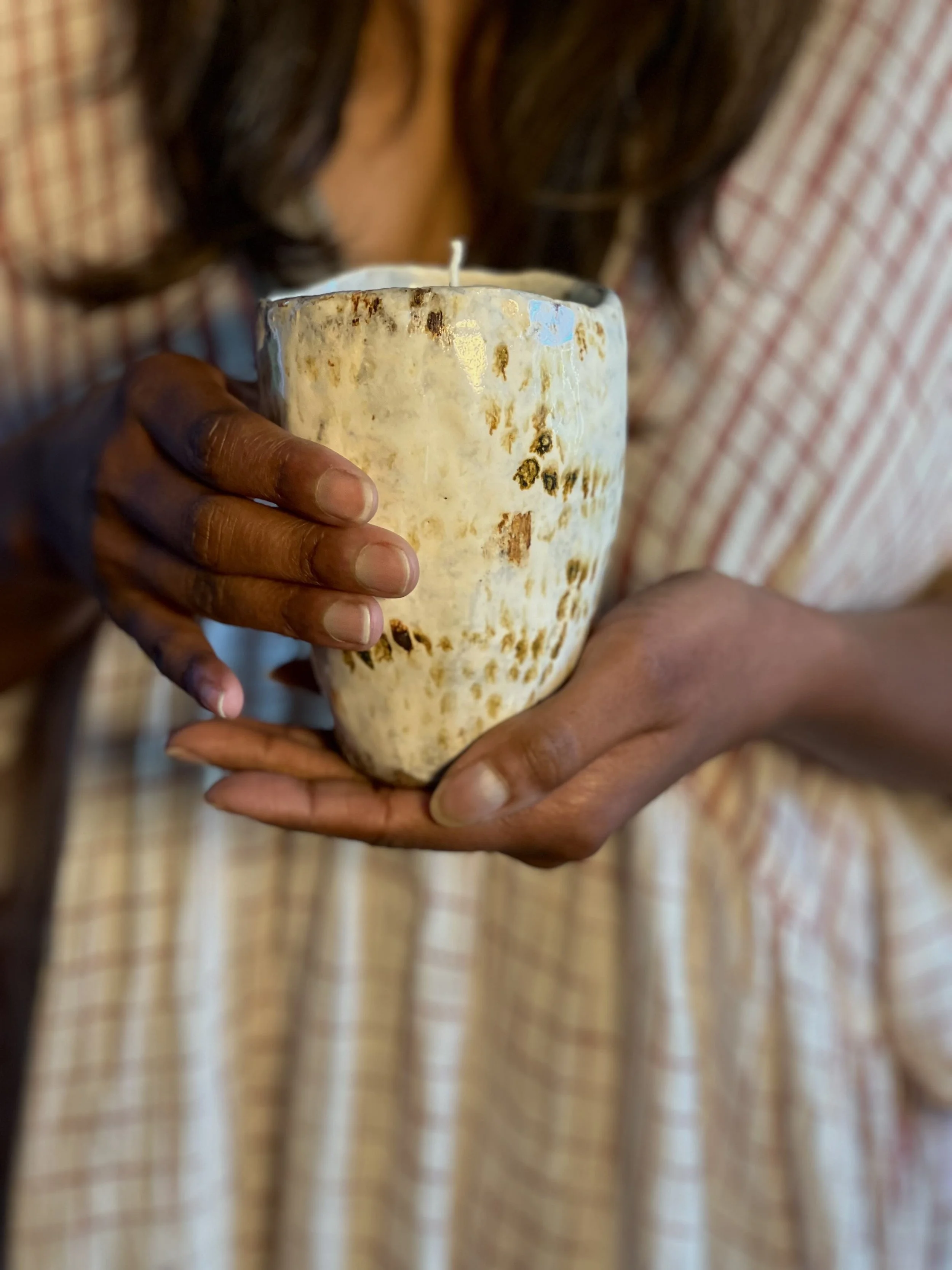 Person holding a large, textured candle in their hands, with a blurred background of a striped fabric garment.